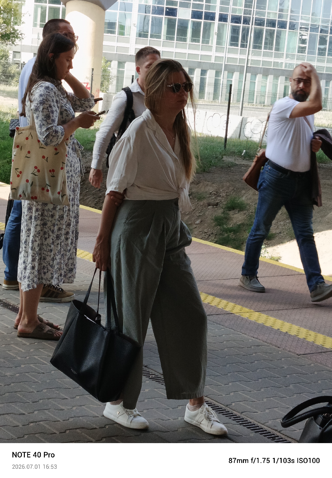The media file captures a group of people standing and walking on a paved area, possibly a train station platform, in Warsaw, Poland. The scene appears to be set outdoors during the daytime, with ample natural light suggesting fair weather.
In the foreground, a woman in a white button-up shirt tied at the waist and loose-fitting grey pants walks towards the right of the frame. She is carrying a large black handbag and wearing white sneakers. To her left, another woman in a blue and white floral dress is seen from the waist down, also holding a light-colored tote bag adorned with cherries. Her feet are clad in sandals and a sneaker. Further back, a man in a white t-shirt and jeans walks away from the camera, with a brown bag slung over his shoulder. Another man, partially visible, is also present.
The background features a modern building with a facade of large glass windows, suggesting an urban or commercial setting. A black metal fence separates the paved area from a grassy, unkempt embankment behind it. The ground is paved with interlocking stones, and a yellow tactile paving strip runs along the edge of the platform.
The timestamp on the media file indicates July 1, 2026, at 16:53, and camera settings of 87mm f/1.75 1/103s ISO100. The text "NOTE 40 Pro" is also visible, likely referring to the device used to capture the image. The overall atmosphere seems casual, possibly a commute or a brief stop during travel.