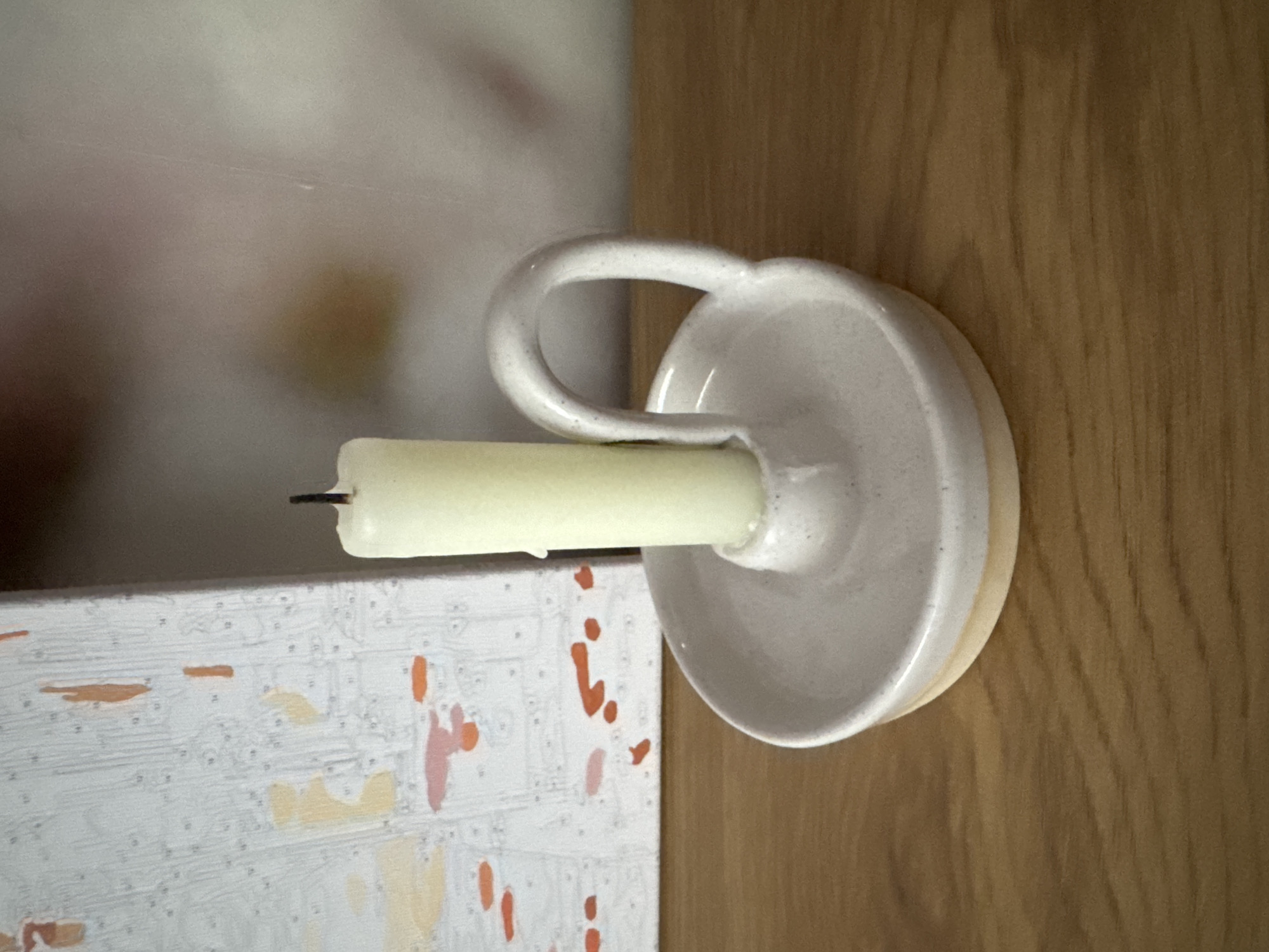 A cream-colored candle is held upright in a white ceramic candle holder. The candle holder is shallow with a wide base and a small, circular depression in the center for the candle. A loop handle is attached to one side of the candle holder. The candle holder is placed on a wooden surface. To the left of the candle holder is a portion of a framed canvas with a paint-by-numbers design, featuring abstract shapes in orange and pink tones on a white background. The background behind the candle holder is blurred and light-colored. The scene is indoors.