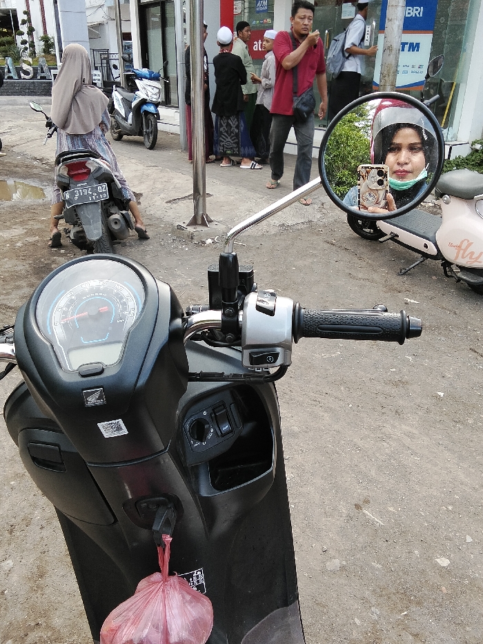 This image depicts a street scene in Pajarakan Kulon, Indonesia. In the immediate foreground, the handlebars and dashboard of a black Honda automatic scooter are visible. The scooter's right-side rearview mirror reflects the face of a woman wearing a maroon helmet, a black head covering, and a light green face mask pulled down to her chin. She is holding a light-colored smartphone with a decorative case, seemingly capturing her reflection. A small, red plastic bag hangs from the ignition key of the black scooter.
In the midground, a woman in a light brown hijab and a patterned dress is seated on a grey scooter (license plate N 3194 OZ), facing away from the viewer. Her feet are on the ground, wearing flip-flops. Behind her, a blue and white scooter is parked. To the right of the seated woman, a group of approximately six men stands on a sidewalk. One man in a red shirt and dark pants, carrying a black shoulder bag, is prominent, appearing to be talking or eating. Several other men wear traditional Muslim attire. A metal pole stands vertically in this area. Puddles of water are present on the uneven, dirty ground.
In the background, a light-colored building with "ATM BRI" signage is visible, indicating a bank. Another building with "ASSA" signage is partially visible on the far left. A light pink electric scooter is partially visible on the far right of the frame, near the reflection in the mirror.