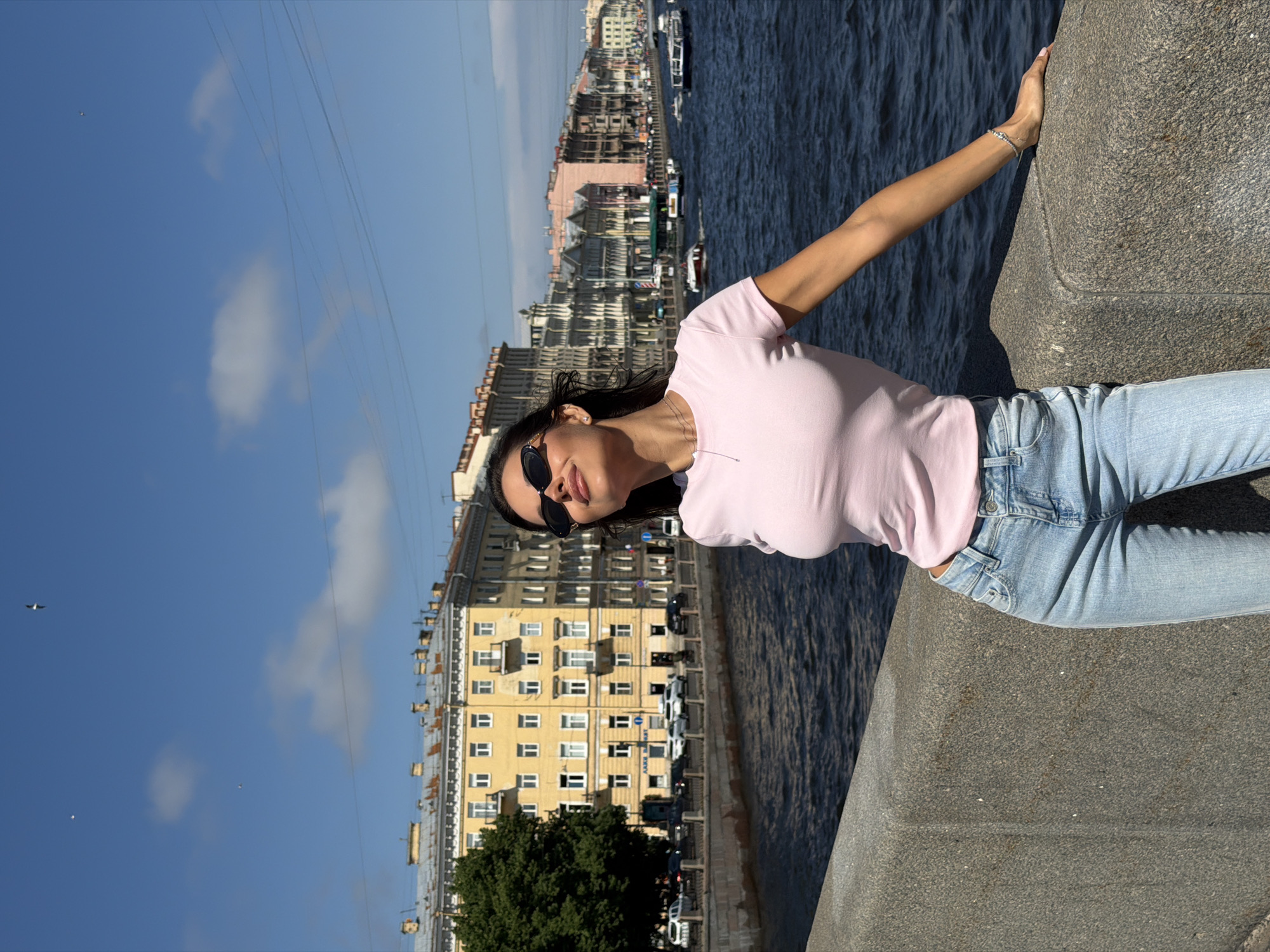 A woman stands on a stone embankment, her body angled towards the right of the frame. She is wearing a light pink t-shirt and light blue jeans, with sunglasses perched on her nose. Her head is tilted back, facing the sky, and her eyes are closed, basking in the sunlight. To her left, a dark blue river flows, with several boats dotting its surface. Across the river, a row of buildings with yellow and pink facades line the embankment, characteristic of Saint Petersburg, Russia. The sky above is a clear blue with a few wispy clouds and two distant birds. The overall scene depicts a bright, sunny day in the historic city.