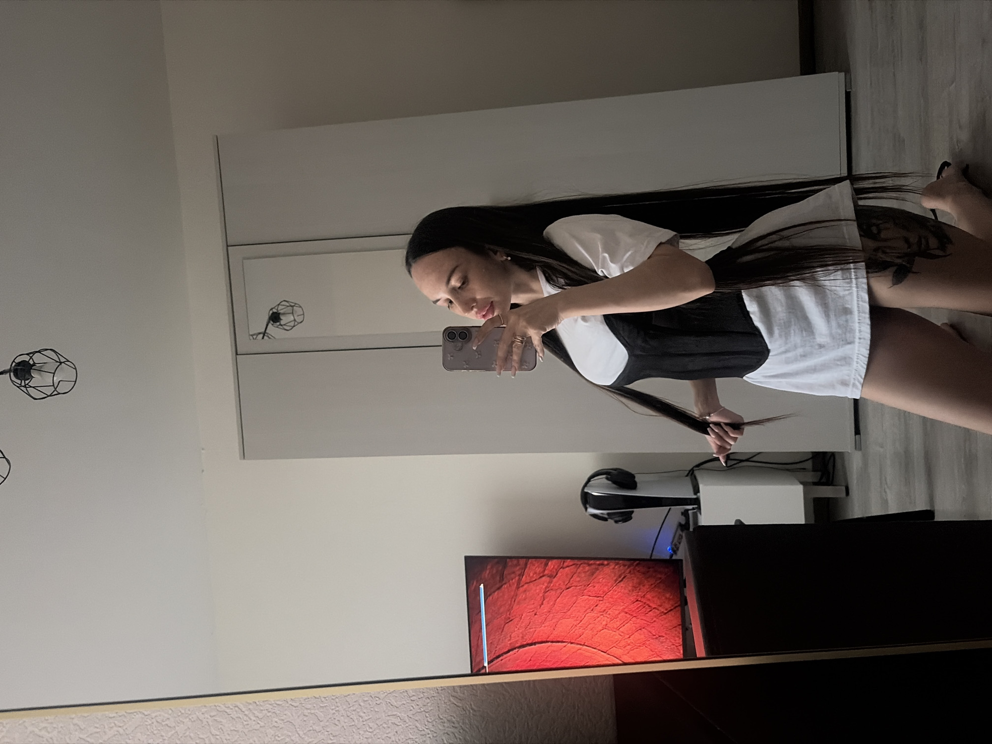 A woman is taking a selfie in a mirror, likely indoors. She is wearing a white shirt and a black corset. Her hair is long and dark. She is holding a phone in her hand. The background includes a white wardrobe, a computer monitor with a red screen, headphones, and a white box. The location is likely a private residence and there's no indication that this is in the city of Saint Petersburg, Russia.