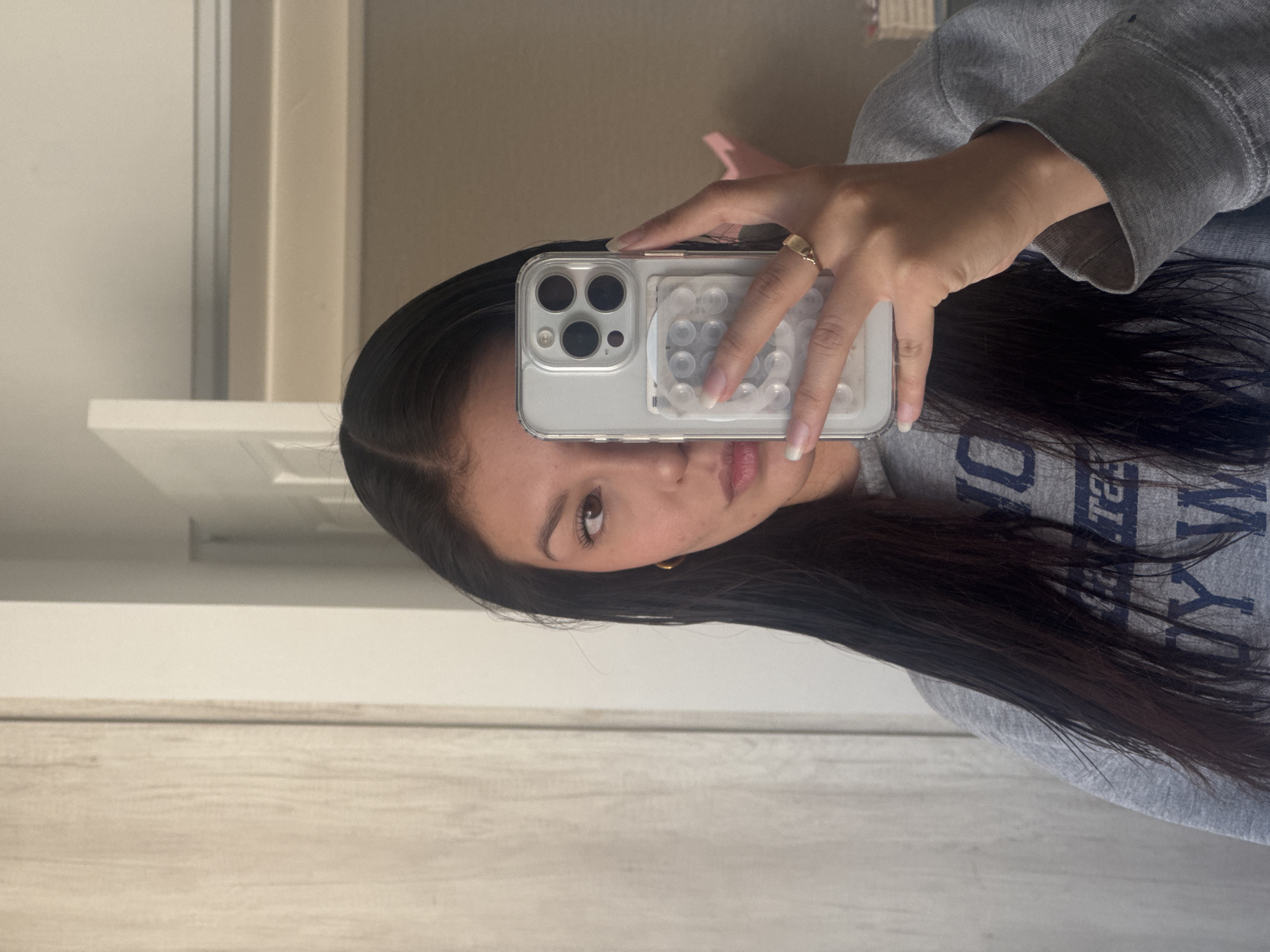 A person is holding a silver iPhone in a clear case with circular grips on the back. The person is wearing a light gray sweatshirt and is taking a selfie in front of a white door and light tan wall. The person has long, dark hair and is wearing gold hoop earrings. A ring is visible on their left hand. The location is not identifiable as San Francisco Ocotlán (Ocotlán), Mexico from this image.
