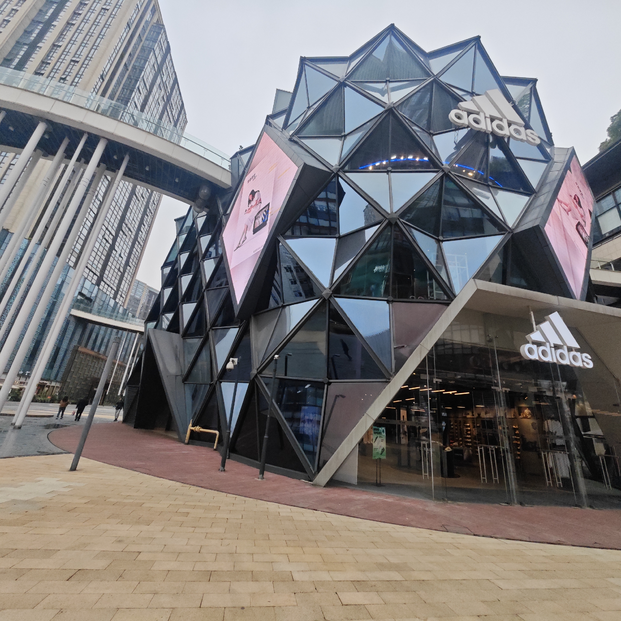 A ground-level shot shows the Adidas flagship store in Yuzhong District, China. The store’s architecture features a geometric facade composed of numerous triangular glass panels, some of which display large digital screens. The Adidas logo is prominently displayed on the building's upper section and above the store entrance.
A tall building with a series of white cylindrical pillars and a connecting skywalk is visible in the background. Pedestrians are walking in the plaza in front of the store, which is paved with light brown brick. The plaza surface is bordered by a reddish-brown surface along the building. The sky is overcast.