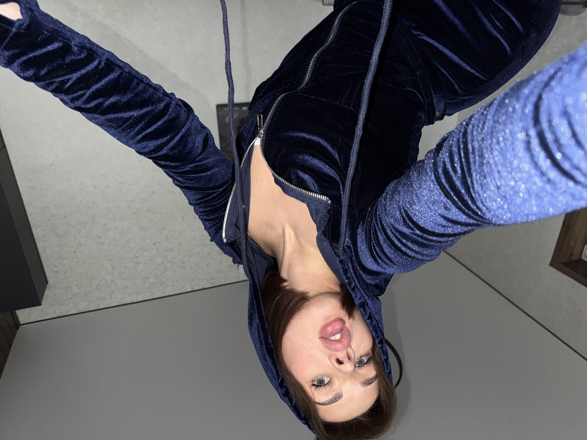 A woman is taking a selfie in what appears to be an indoor setting. She is wearing a dark blue, possibly velvet, hooded tracksuit with the zipper partially open. Her arms are raised, stretching the sleeves of the tracksuit. The background consists of gray flooring and a section of a black cabinet or shelf to the left, and a light-colored wall with a wooden framed object to the right. There is no indication of the location being Лобаново, Russia, from the image itself.