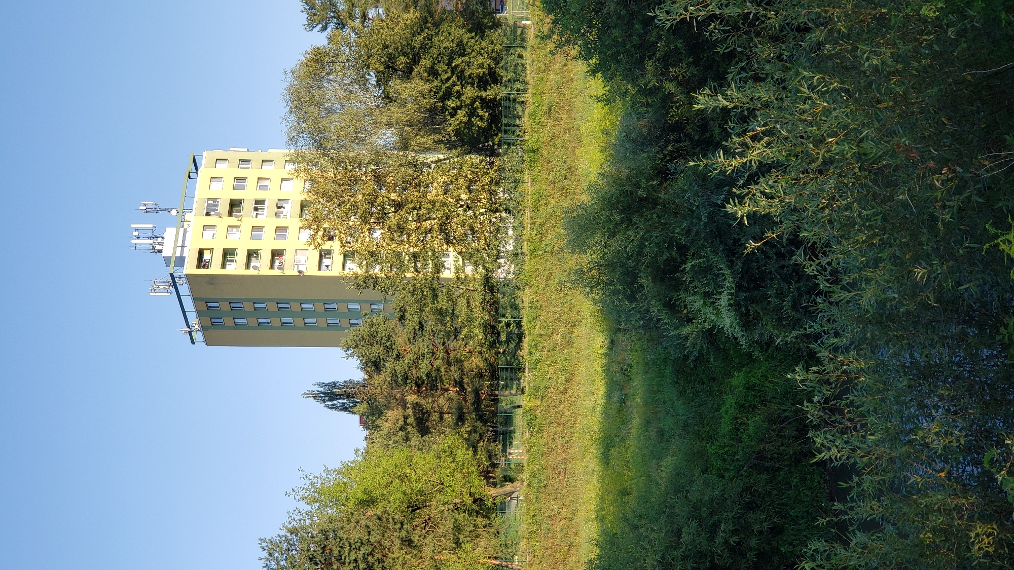 The image shows a tall, yellow and green apartment building with several antennas on its roof. The building is surrounded by trees and a grassy area. The sky is clear and blue. The apartment building is likely located in the city of Prešov, Slovakia. The building appears to be a typical panelák, a common type of housing estate apartment building in socialist countries. The antennas on the roof are likely for mobile phone communication or broadcasting. The area around the building is green and lush, suggesting it is a residential area with parks or green spaces. There are no people visible in the image.