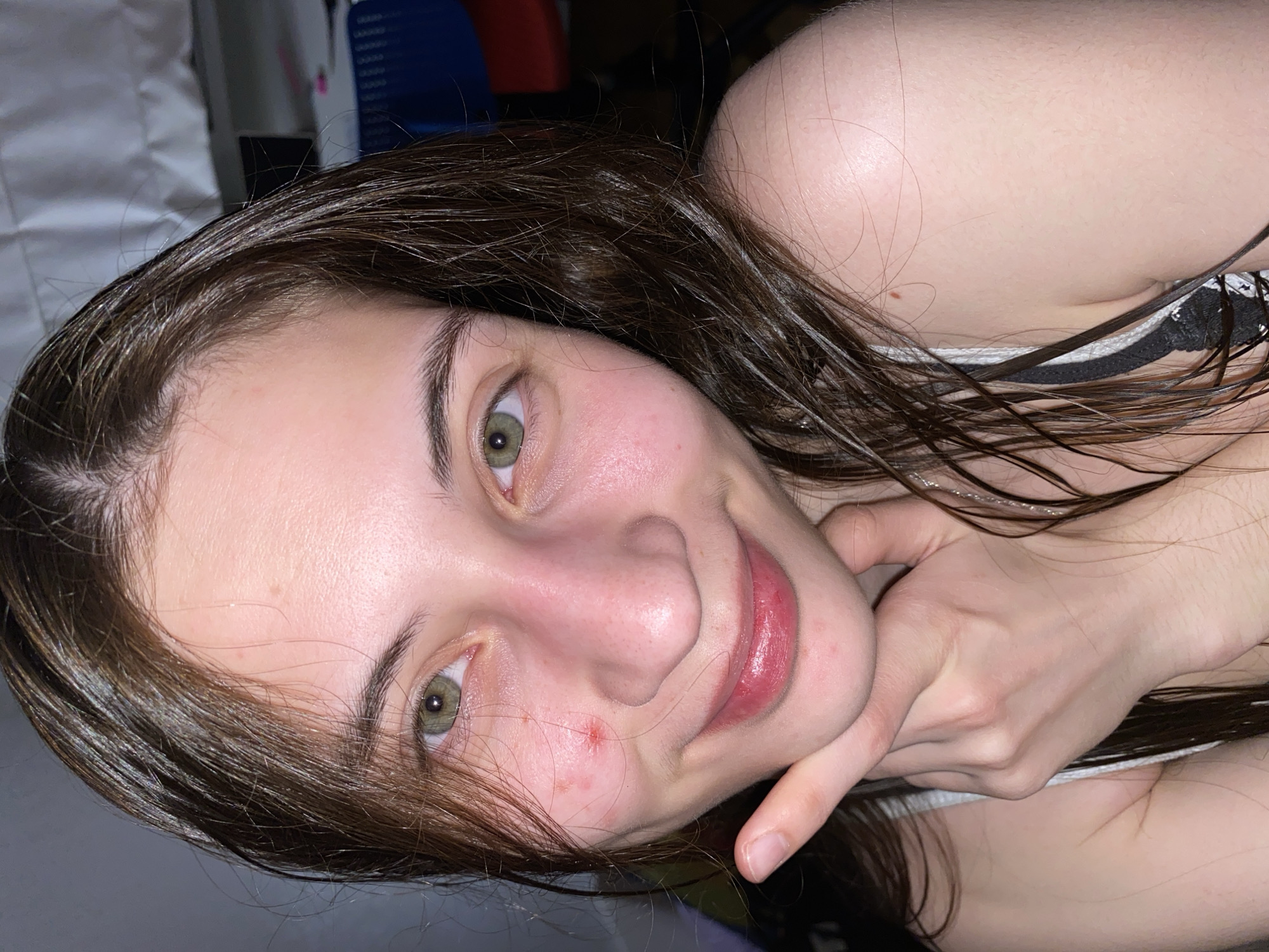 A young woman is seen in a close-up, slightly rotated view. Her dark, wet hair is partially covering her shoulders and face. She has green eyes and fair skin. There are some blemishes visible on her face. She is resting her chin on her index finger. Part of a striped garment is visible on her shoulder. The background is indistinct, showing blurry elements of what appears to be a room. The location cannot be determined from the image. There is no evidence to suggest this photo was taken in Votkinsk, Russia.