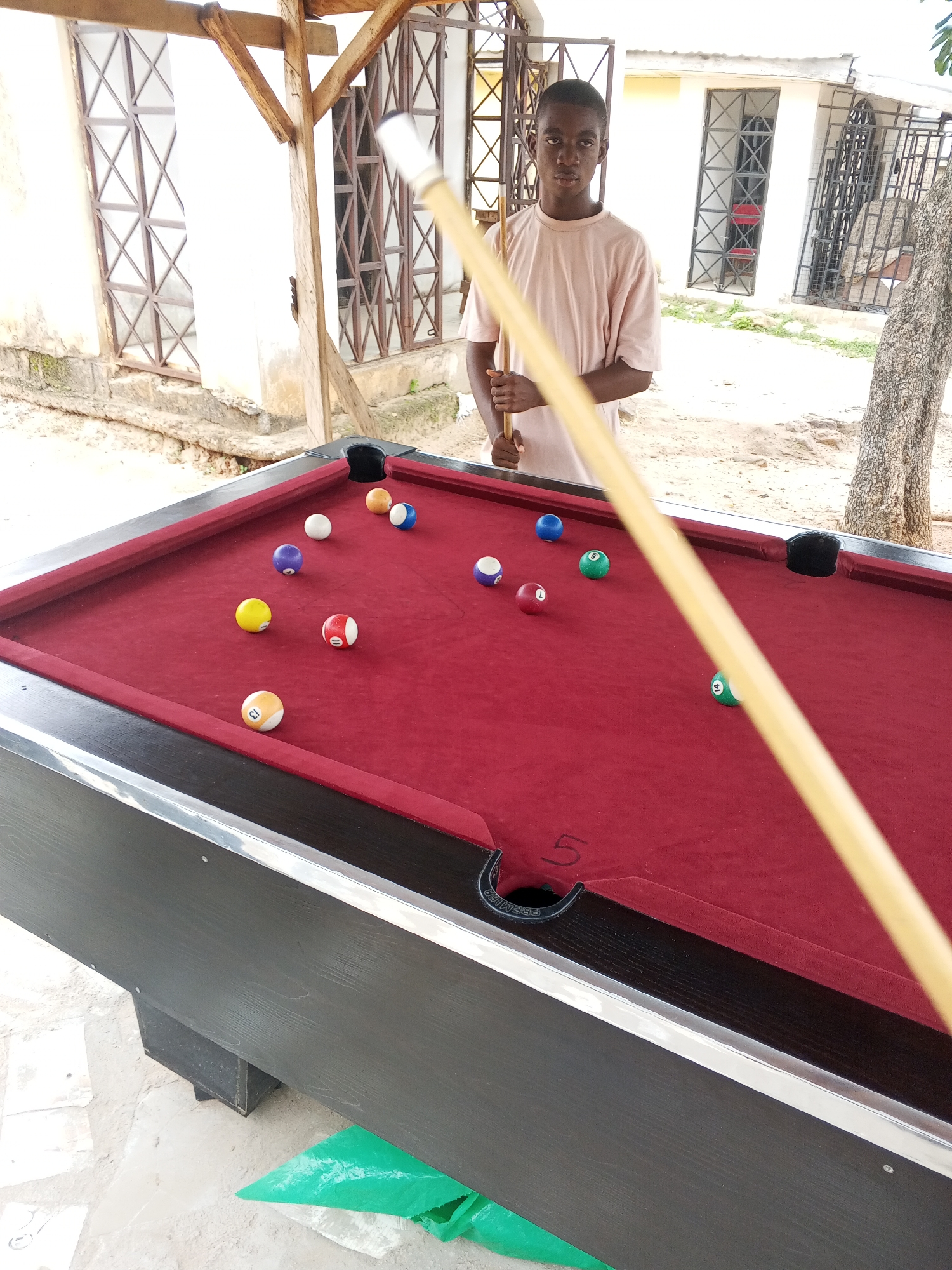 A young Black male, appearing to be in his late teens or early twenties, stands behind a red-felt billiard table, holding a cue stick. He is wearing a light-colored t-shirt. The billiard table has several colored balls scattered across its surface, including a yellow ball with the number 13, a red and white ball with the number 5, a purple ball with the number 3, a green ball with the number 9, a blue ball with the number 1, a green ball with the number 14, and a blue ball. The scene is outdoors, indicated by the natural light, a tree trunk to the right, and a rough, sandy ground. In the background, there is a building with white walls and barred windows, suggestive of a courtyard or an open-air structure. The overall impression is of a casual setting for playing pool. The location is identified as Lokoja, Nigeria.