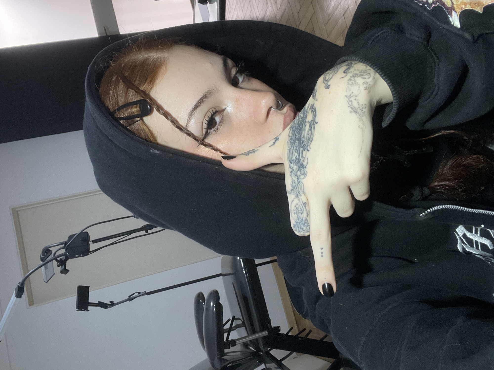 A young woman with fair skin, freckles, and brown hair partially tucked under a black hooded sweatshirt poses for a low-angle selfie indoors. She has a nose ring and visible tattoos on her right hand. Her nails are painted black, and she's pointing downwards with her right index finger. A black hair clip is visible in her hair near her left ear. Behind her, there is a chair and a lamp fixture. The location is unidentified; there is no verifiable evidence in the image to place it in Buenos Aires, Argentina.