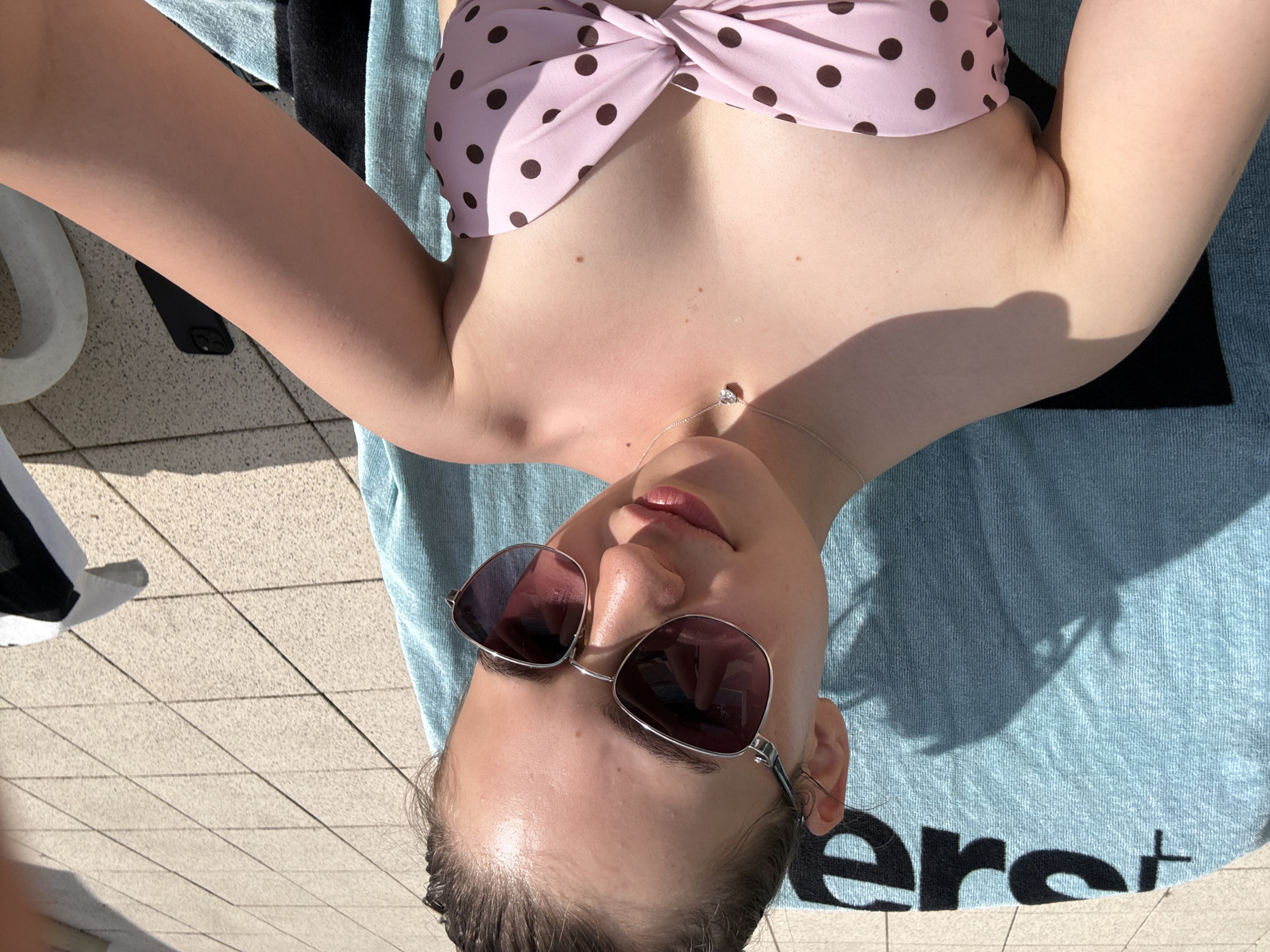 The image shows a young woman in a pink polka-dot bikini top and sunglasses, relaxing on a blue towel outdoors. She is looking at the camera and appears to be taking a selfie. The background consists of a paved patio area. There is a black phone on the ground near her. There is no indication of the location being the city of Forbach, Germany.