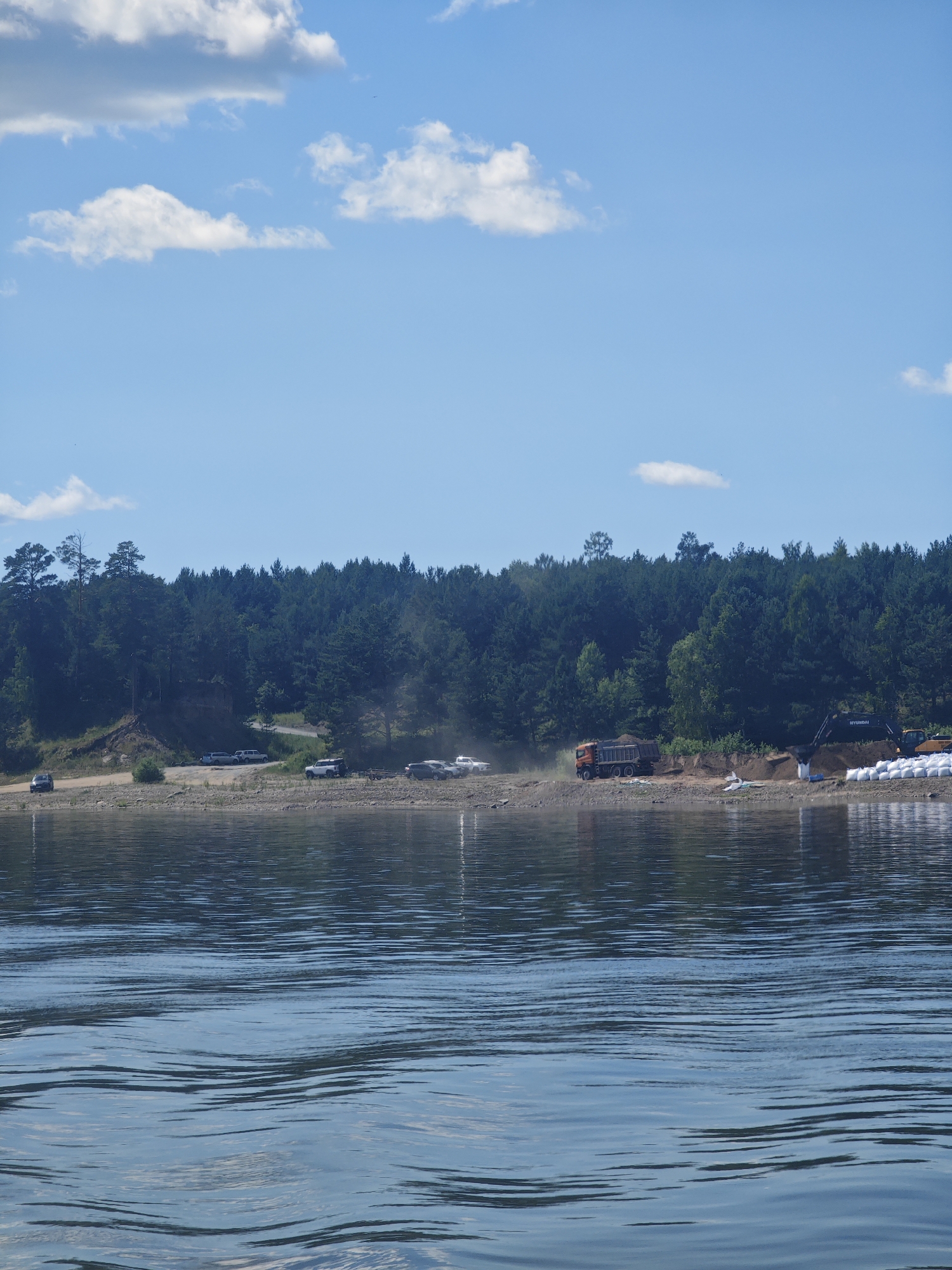 The image depicts a waterfront scene on a sunny day, likely in Strelka, Russia, given the context. The foreground is dominated by rippling water, reflecting the sky and the distant shore. The middle ground features a rocky bank bordering a dense forest of evergreen trees that stretches across the horizon. Along the bank, several vehicles are parked, including cars and what appears to be a truck. Evidence of construction or land reclamation is visible, with a large orange dump truck, an excavator, and rows of white sandbags or similar materials stacked on the shore. The sky is a vibrant blue with scattered white clouds.