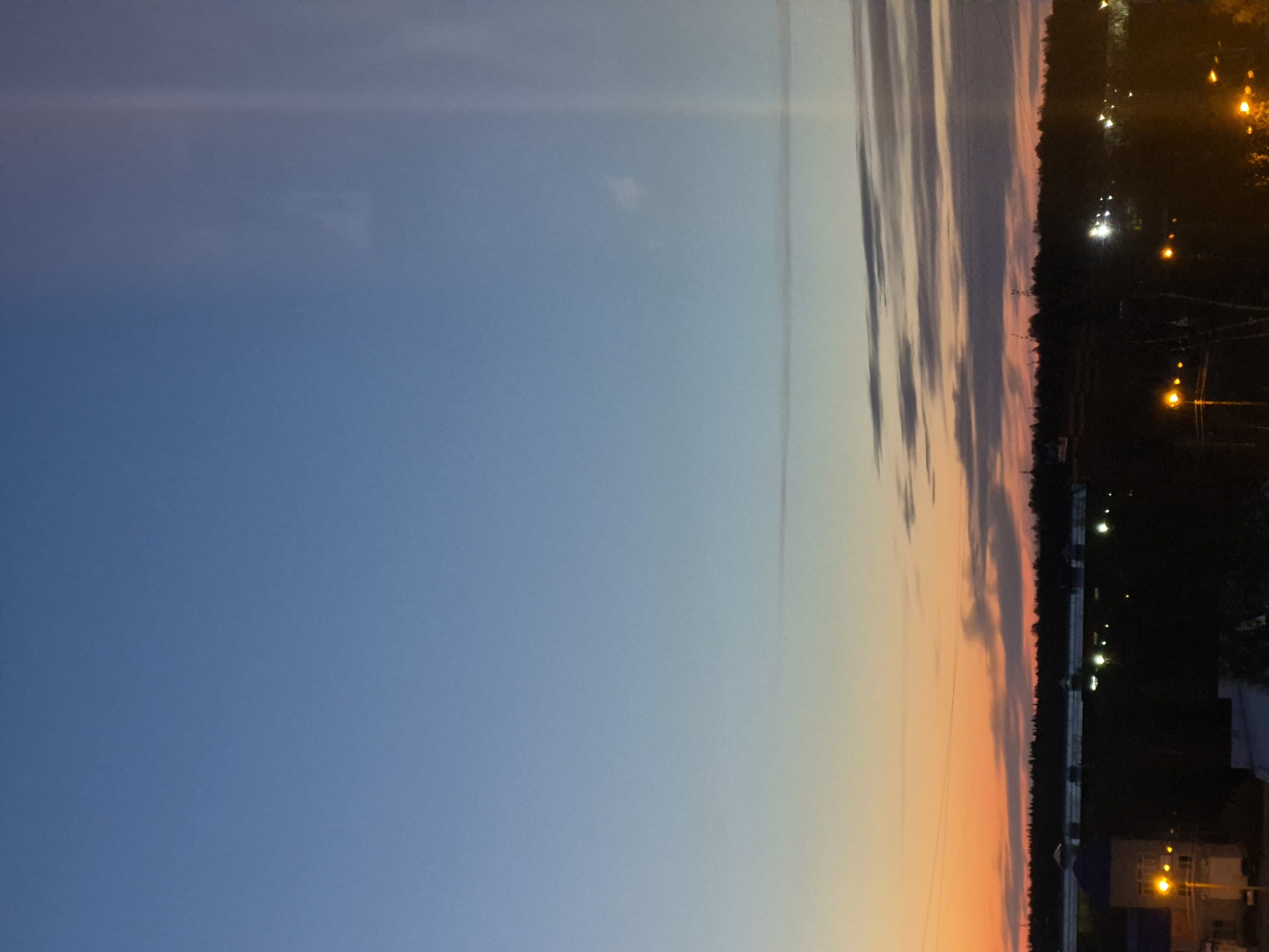 The image shows a sunset sky over Tobolsk, Russia. The sky transitions from a deep blue on the left to orange and pink hues near the horizon. There are some clouds and streaks visible in the sky. The horizon line is lined with trees and buildings, with streetlights illuminating the area. The scene is dark at the bottom edge where the city is located.