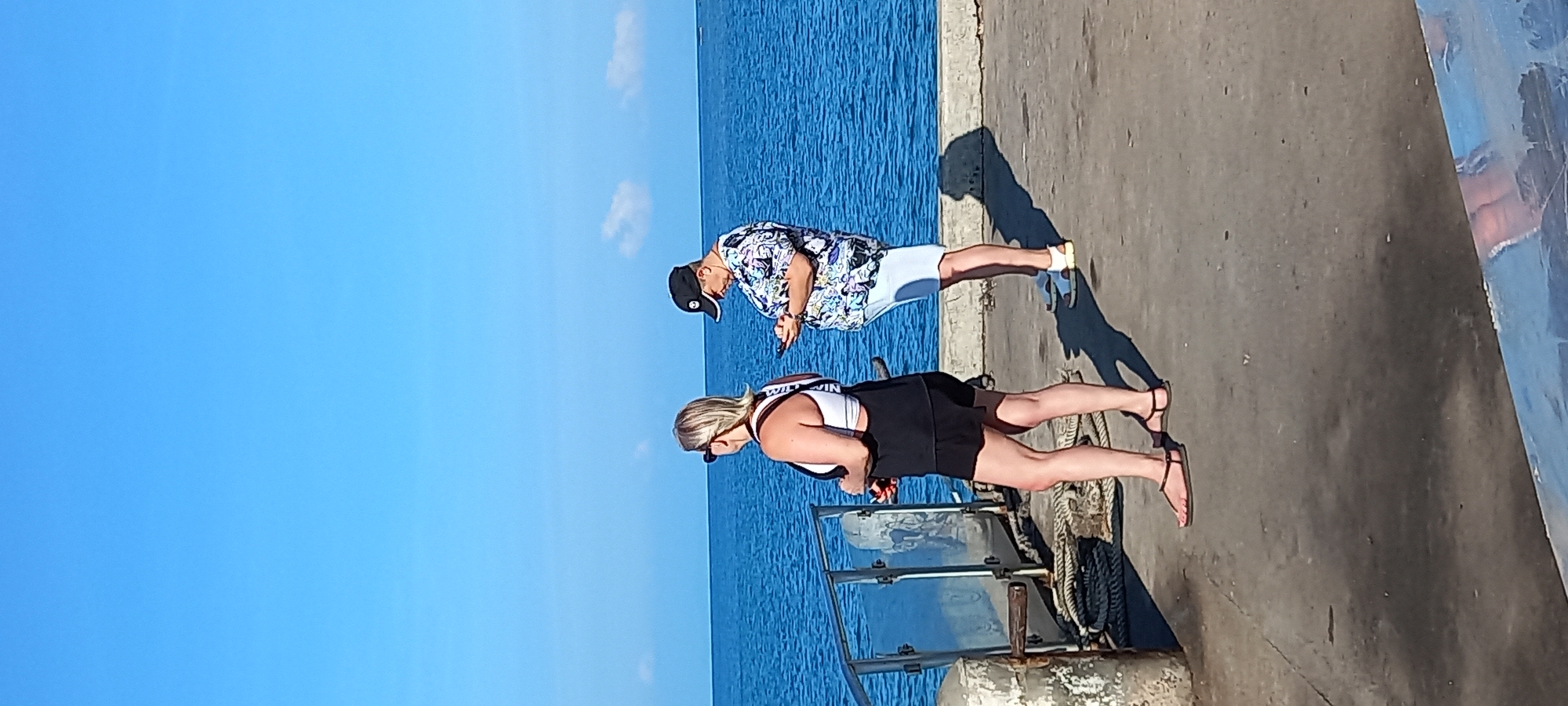 This image depicts a sunny outdoor scene at a waterfront location, likely a pier or dock in Thong Sala, Thailand. The sky above is a clear, bright blue, meeting a vivid blue body of water.
Two individuals are standing on a concrete pier. Closer to the camera, a person with blonde hair, wearing a white sports bra-style top with black straps, black shorts, and dark sandals, stands near a transparent barrier with metal framing. The text "WILD WIN" is faintly visible on the black strap of their top. This individual is wearing sunglasses and appears to be looking downwards or away.
Further back on the pier, another person is visible, wearing a patterned short-sleeved shirt (featuring blue, white, and black designs), light-colored shorts, and a black baseball cap worn backward. This person is holding a smartphone and looking down at it, seemingly engrossed in its contents.
The strong sunlight creates distinct shadows of both individuals on the concrete pier, suggesting it is mid-day to early afternoon. Coiled ropes or mooring lines are visible on the pier near the transparent barrier. A large blue structure or part of a boat is partially seen on the far right edge of the frame. The overall atmosphere is bright and relaxed, indicative of a warm, coastal environment.