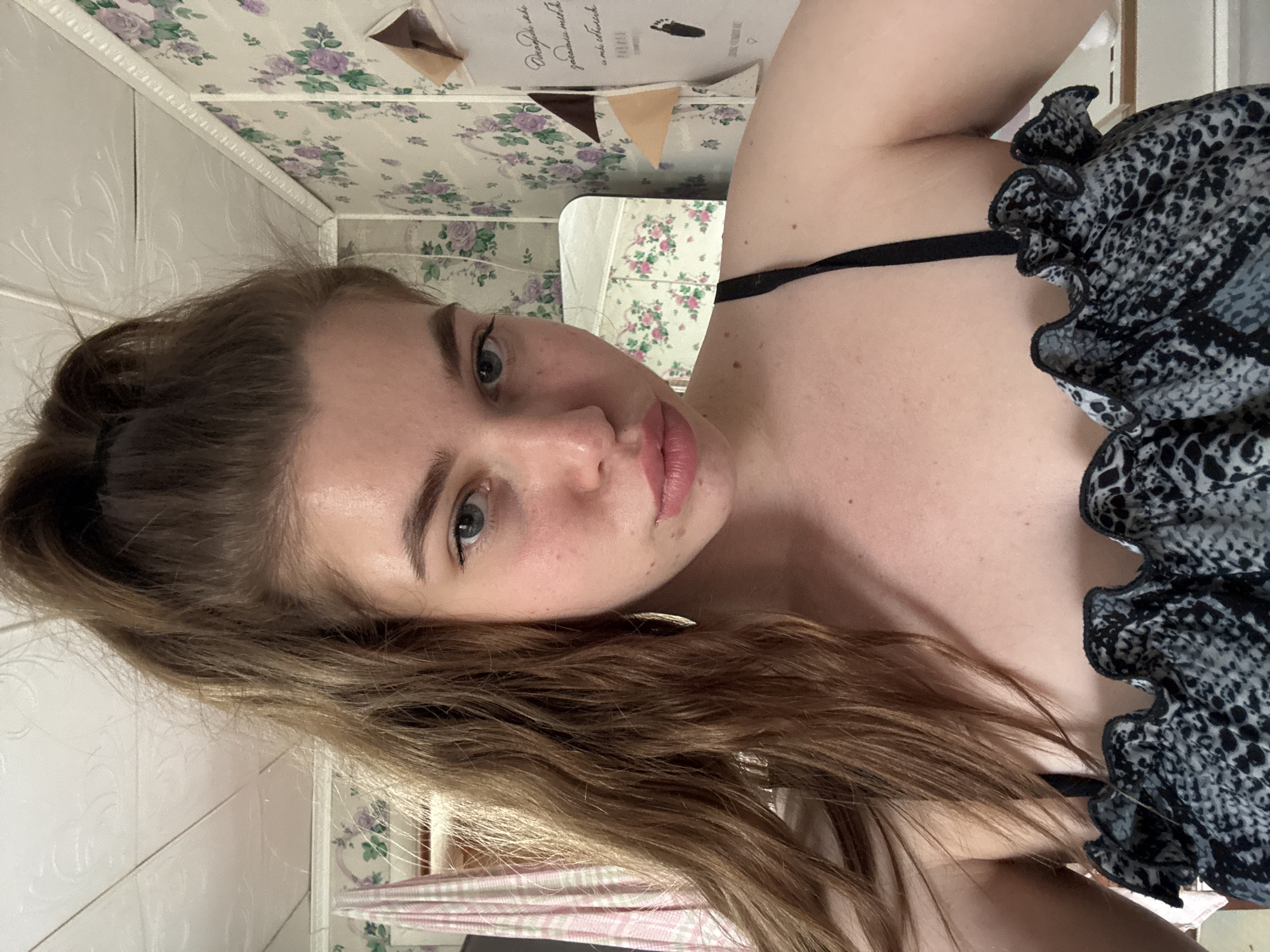 A young woman with fair skin, long light brown hair pulled back, and blue eyes is posing for a selfie. She is wearing a black and gray patterned top with ruffled detailing and thin black straps. The background features a bathroom with a floral patterned wallpaper and tiled walls. A mirror reflects more of the floral pattern and some indistinct objects. The location of Мещёры, Russia, is not visually verifiable from this image.