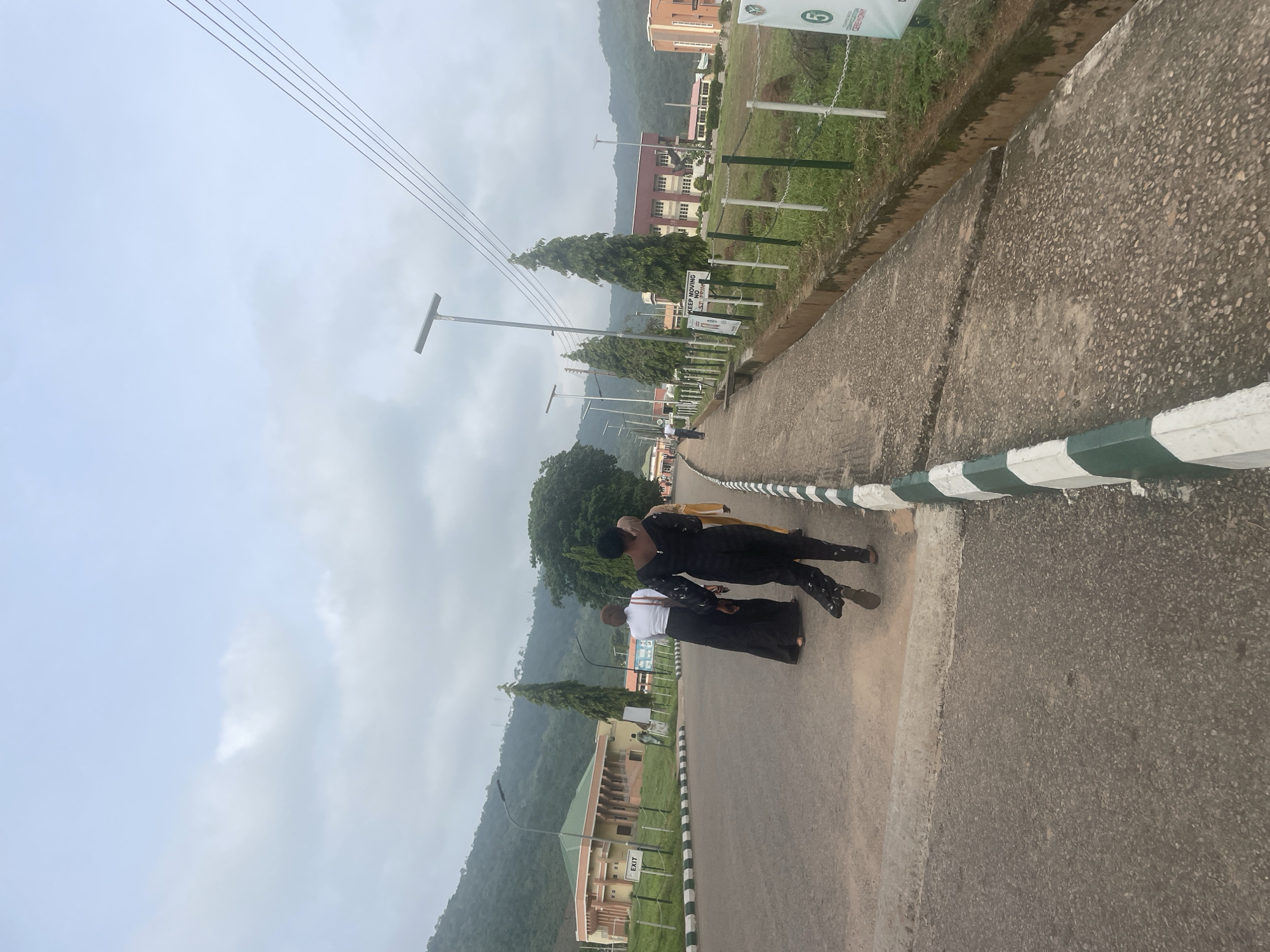 The image captures an outdoor scene on a cloudy day, likely on a university campus in Lokoja, Nigeria, given the architectural style and surrounding hilly terrain. In the foreground, a paved pathway with a green and white striped curb runs along a grassy area. Several people are walking along the pathway. Three individuals are prominently visible in the middle ground, with their backs to the camera. One person is wearing a black outfit with patterned leggings, another a white shirt and black skirt, and a third in a yellow garment. In the background, several buildings with modern architecture, characterized by red bricks and large windows, are situated amidst lush green hills. Power lines and lampposts are also visible. The overall atmosphere suggests a casual daytime setting on an educational institution's grounds.
