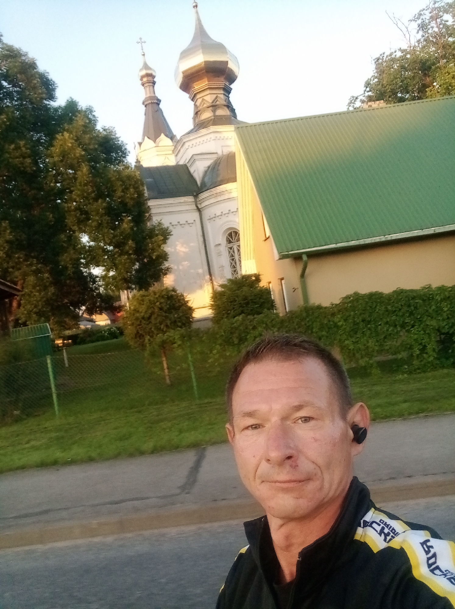 The image shows a selfie of a man with short, graying hair and blue eyes. He is wearing a black jacket with yellow and white stripes. He has a dark earbud in his right ear. Behind him, a church with a silver dome and a green roof is visible. The church is white with decorative trim. Trees and a grassy area with a fence are also in the background. The scene appears to be outdoors in Tapa, Estonia, during the day, possibly in the late afternoon or early evening, given the lighting.