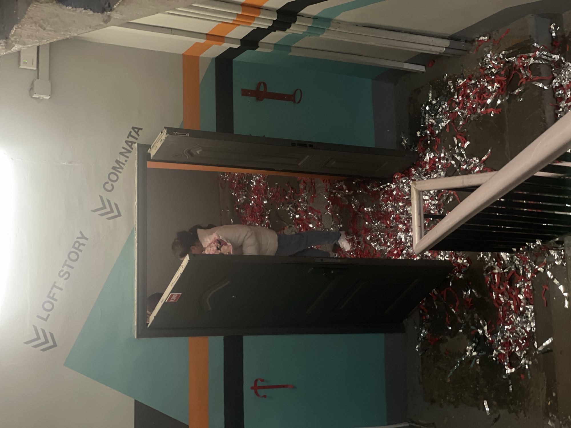 The scene appears to be an interior space, possibly a loft-style apartment or commercial area. The walls are painted with geometric patterns in colors including teal, white, orange, and black. Signage with the phrases "LOFT STORY" and "COM.NATA" is visible.
A doorway is ajar, and a person in a beige jacket and jeans is lying on the floor within the room. Red and silver tinsel or confetti is scattered throughout the room. There's a railing or banister visible on the right side of the image.
A red object that looks like a coat hook or hanger is on the wall above the doorway.
Based on the information provided, this is likely the "Loft Story Comnata" in Saint Petersburg, Russia.