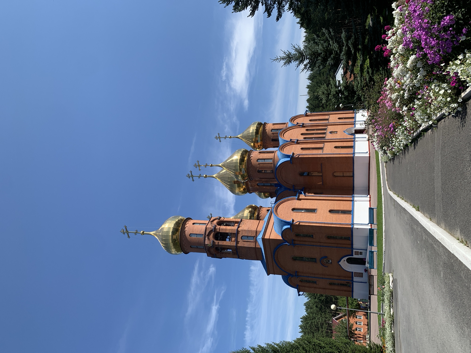 A bright, wide-angle, eye-level outdoor shot captures an Orthodox church with multiple golden domes under a clear blue sky with sparse white clouds. The church is constructed of red brick with blue trim and white detailing. A prominent bell tower stands to the left of the main structure, also adorned with a golden dome. Several other golden domes rise from the church roof, each topped with a cross.
In the foreground, an asphalt pathway curves to the right, bordered by a vibrant flowerbed filled with an array of pink, white, and purple flowers. To the left of the path, a paved walkway leads towards the church entrance. Beyond the pathway, a strip of green grass separates it from the church's white foundation. A few dark benches are placed on the grass in front of the church.
To the far left, a cluster of pine trees stands tall. Behind the church, a dense forest of evergreen trees forms the backdrop. In the mid-ground, to the left of the church, a smaller brick building with a red roof and white window frames is visible, with a lamppost nearby. Two figures can be seen walking near this building.
The scene is peaceful and well-maintained, suggesting a serene environment. This church is identified as the Church of the Resurrection of Christ in the city of Prokopyevsk, Russia.