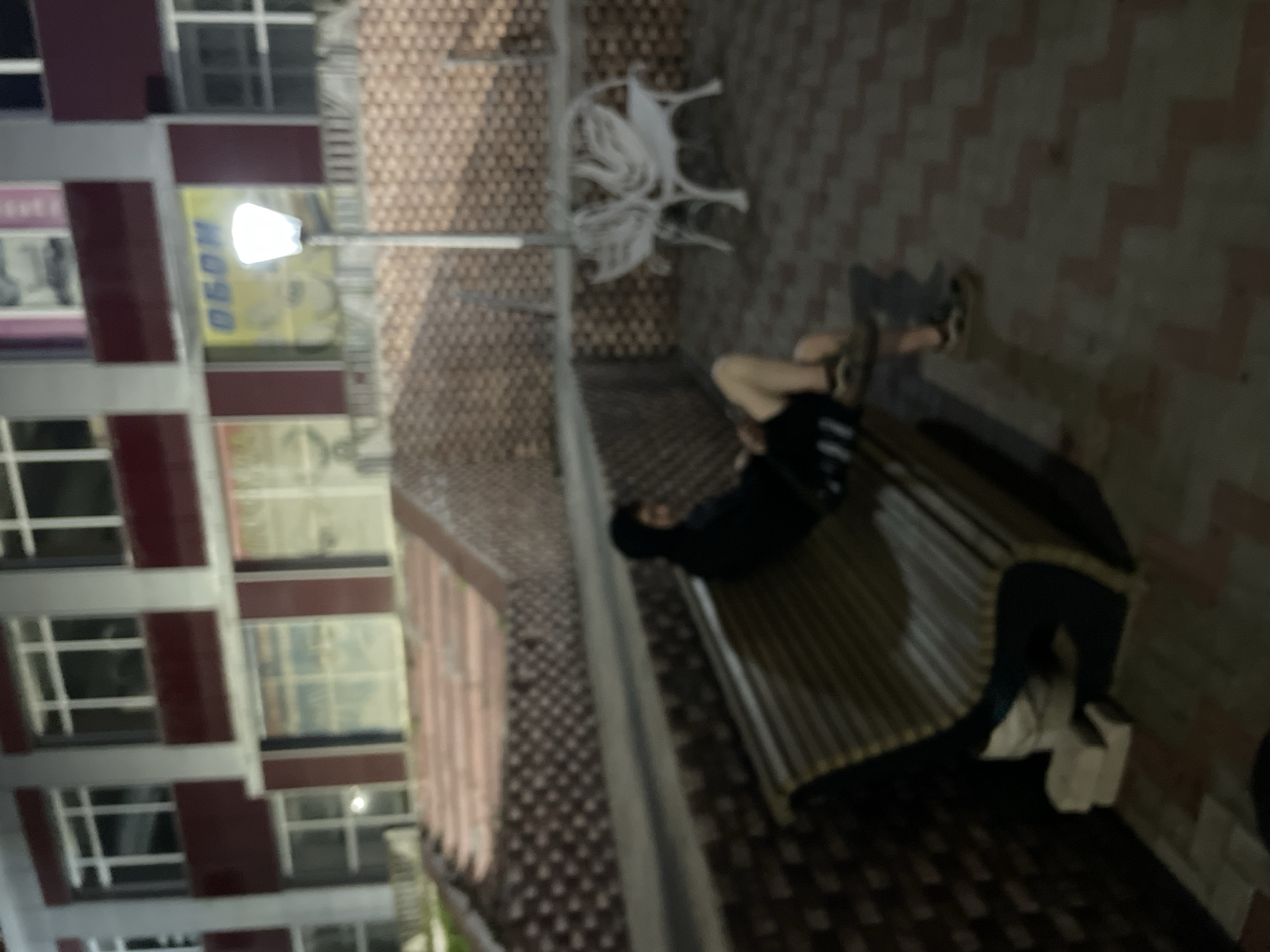 A person wearing a black hoodie and shorts is sitting on a wooden bench in front of a brick wall. The person is facing to the right of the frame and their legs are extended. The bench is positioned on a tiled patio. In the background, there is a building with windows and a lamppost. The scene appears to be in an outdoor urban setting, likely in the city of Krasnye Baki, Russia, given the provided context. The lighting suggests it is evening or nighttime.