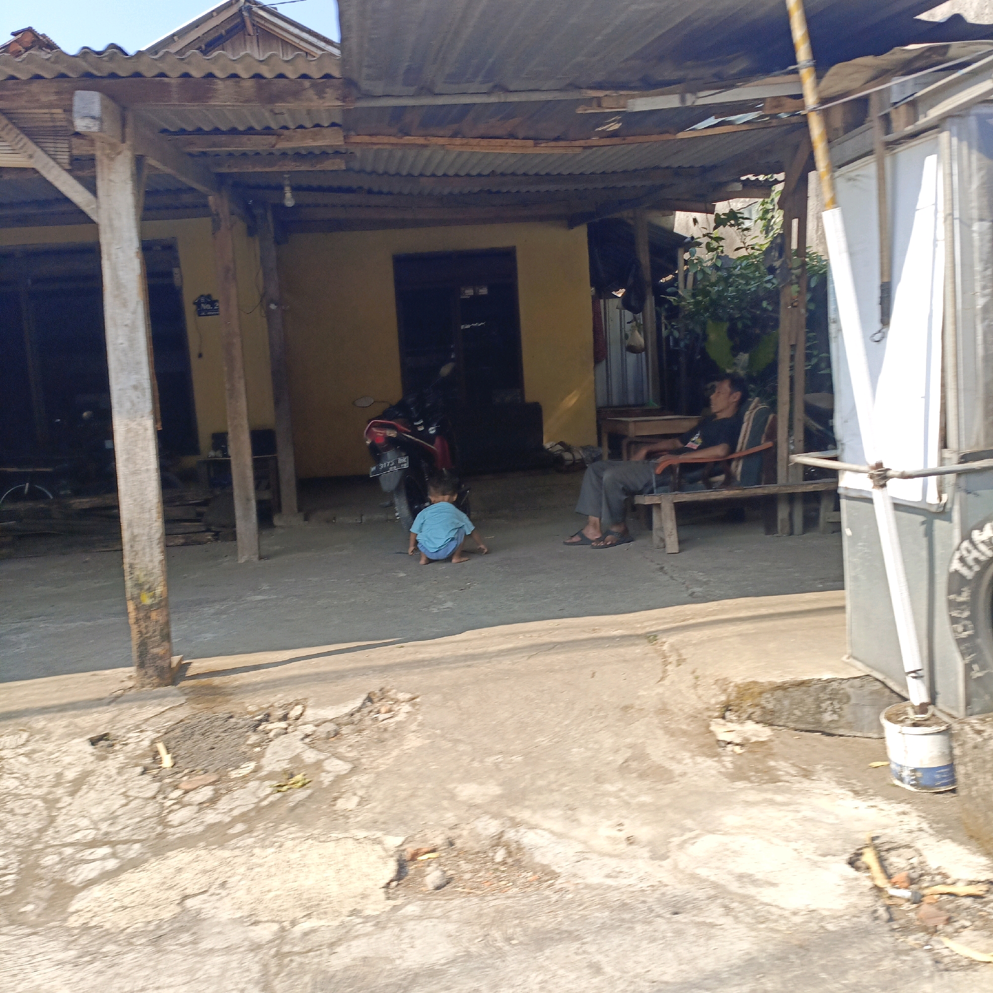 The image depicts an outdoor scene in Salatiga, Indonesia, likely a residential area. In the foreground, a concrete ground shows signs of wear and tear, with scattered debris and shadows cast by overhead structures. A young child wearing a light blue t-shirt and blue shorts is crouched on the ground, facing away from the camera, engaged in play near a red and black motorcycle. To the right, an adult male, dressed in a black t-shirt and dark pants, is seated on a wooden bench, looking to the left. Behind them is a simple building with a corrugated metal roof supported by wooden beams. A motorcycle is parked near the building. The environment appears somewhat unkempt, with visible vegetation in the background and a general sense of a modest dwelling.