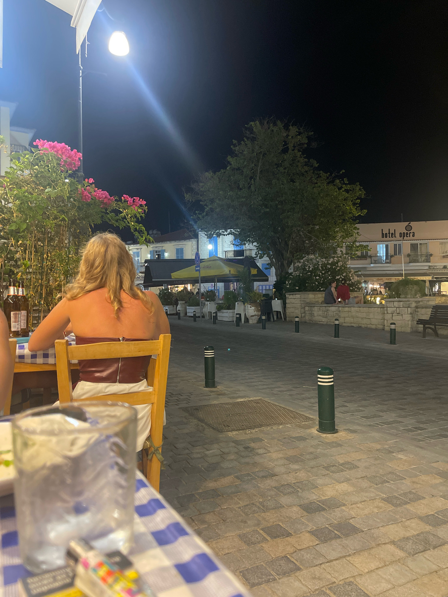 A woman with blonde hair is seated at a table in an outdoor restaurant at night in Larnaca, Cyprus. She is wearing a strapless red top and a white skirt. The table is covered with a blue and white checkered tablecloth. A glass of water and a pack of cigarettes are on the table. In the background, there is a street with paved sidewalks, a tree, and a building with a sign that reads "Hotel Opera." Several other people are visible in the distance, sitting at tables and walking along the street. The scene is illuminated by streetlights and the lights from the hotel and restaurants.