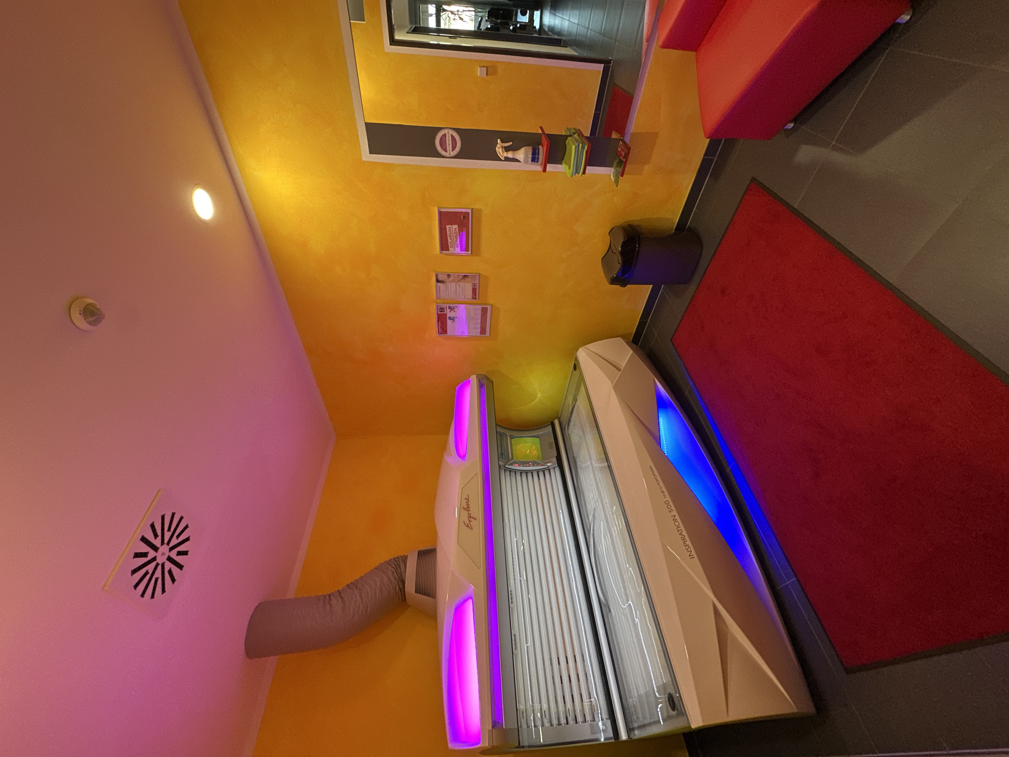 The image depicts the interior of a tanning salon, specifically the "Ergoline" solarium room in Garmisch-Partenkirchen, Germany. The room is brightly lit, with yellow textured walls and a pink-lit ceiling. A large, cream-colored Ergoline Inspiration 500 solarium bed dominates the center of the room, featuring purple and blue LED lighting.
In the foreground, a red mat is placed in front of the solarium bed. To the right, a red sofa is visible. On the yellow wall, there are framed posters, a trash can, and a small shelving unit with cleaning supplies, including a spray bottle and folded towels. A ventilation grate is present on the ceiling, with a flexible duct connected to the solarium. The floor appears to be tiled in a dark gray color. The scene is static, with no people or active interactions. The lighting creates a vibrant and somewhat futuristic atmosphere.