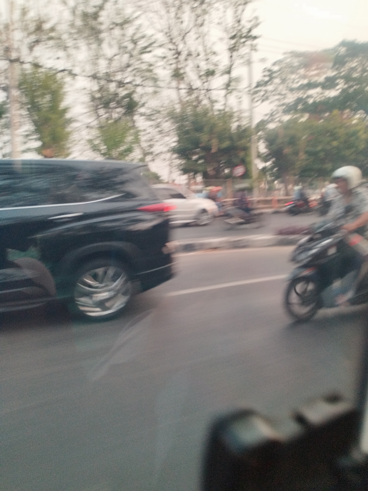 The image depicts a blurry street scene in Surabaya, Indonesia, likely during the day. In the foreground, a black SUV is seen in motion, with its rear wheel clearly visible. The SUV appears to be on a paved road, with lane markings faintly visible. Further down the road, a person on a motorcycle, wearing a white helmet, is also in motion. The background is filled with trees and foliage, characteristic of a city with ample green spaces. The overall impression is one of a busy street with vehicles navigating through traffic.