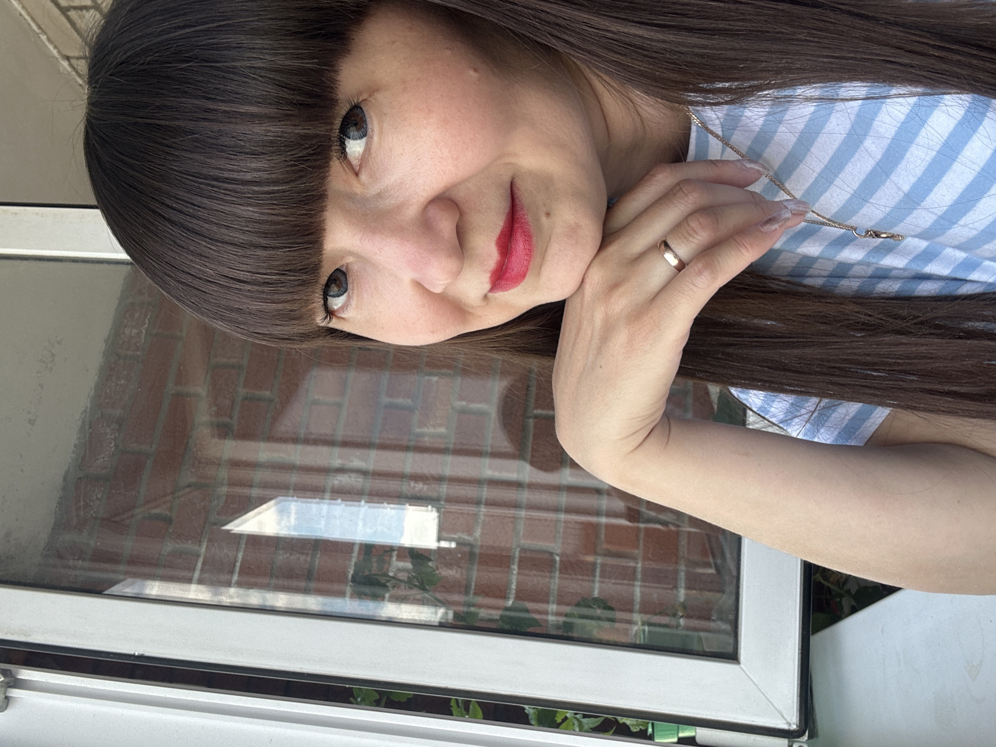 A woman with dark brown hair and bangs is looking at the camera with her head tilted and her chin resting on her hand. She is wearing a blue and white striped shirt and a gold ring on her left hand. Her lips are painted red. She is standing in front of a window with a brick wall visible through the glass. The scene appears to be in a residential area in Chaikovsky, Russia.