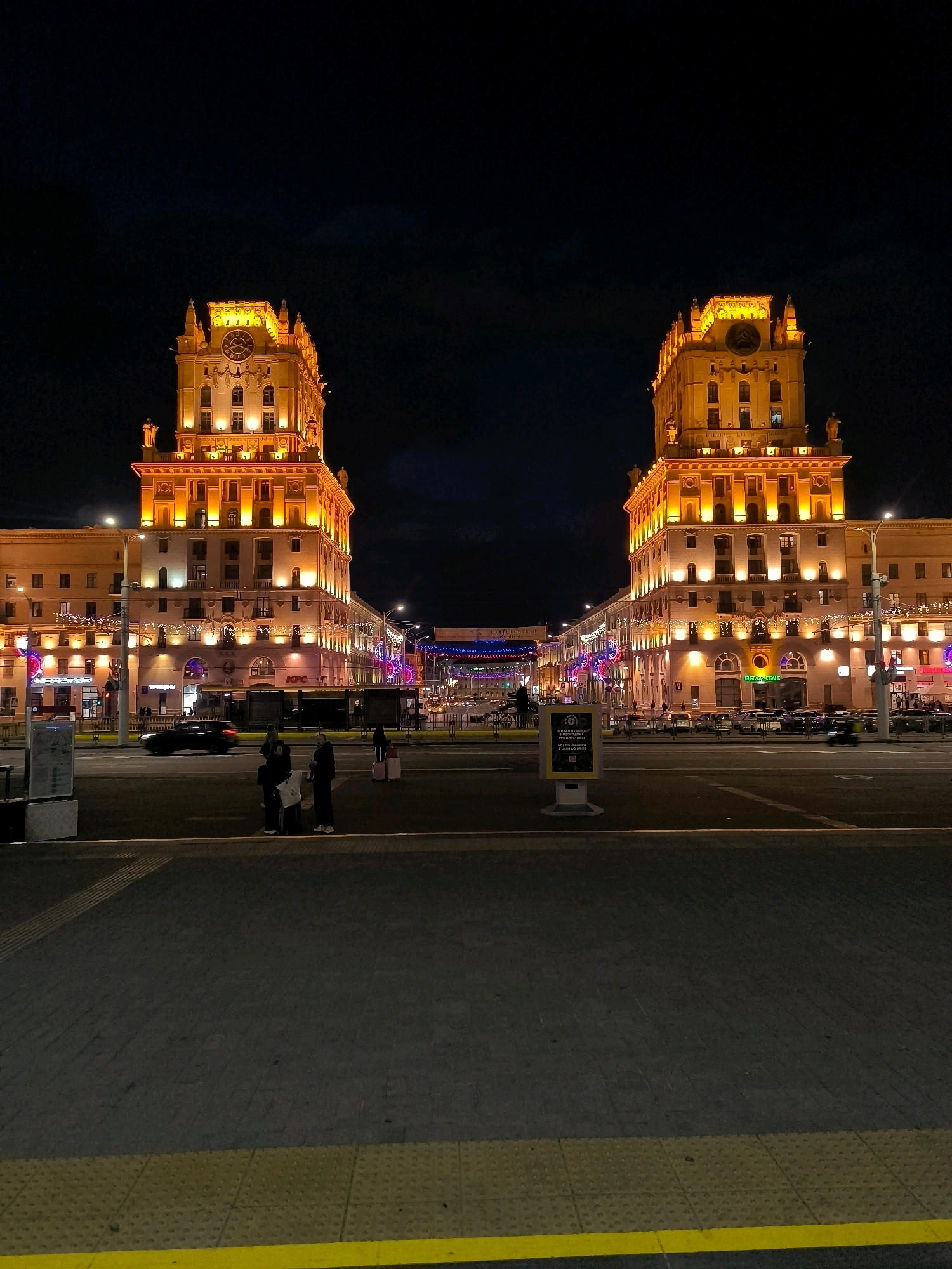 This is a nighttime, eye-level, vertical, full shot of the "Gates of Minsk," also known as the "Stalinsky Towers" or "Vori in Law Towers," located at the entrance to the city of Minsk, Belarus, on Independence Avenue (Praspyekt Nezalezhnastsi). The image showcases the twin, identical Stalinist Empire-style buildings, illuminated by warm, golden lights. The architecture is imposing, with symmetrical facades, ornate detailing, and prominent clock towers on each building. The sky above is dark, with minimal clouds, emphasizing the brightly lit structures.
In the foreground, a paved public space or plaza is visible, with a dark asphalt surface and a contrasting yellow tactile paving strip along the edge. Three individuals are standing on the pavement, facing away from the camera, seemingly waiting or observing. One person, dressed in dark clothing, holds a white bag. Another person, also in dark clothing, stands next to them. A third person, in dark attire, stands further to the right, beside a suitcase. Behind them, a black car is blurred due to motion, indicating it is passing by.
The middle ground reveals a street with several cars and other vehicles, also moving. The buildings lining the street are lit, with storefronts and decorative lights, suggesting a commercial or public area. Further down the avenue, more buildings and streetlights are visible, creating a sense of urban depth. The overall impression is one of a grand, illuminated cityscape at night, with the iconic Stalinist architecture dominating the scene.