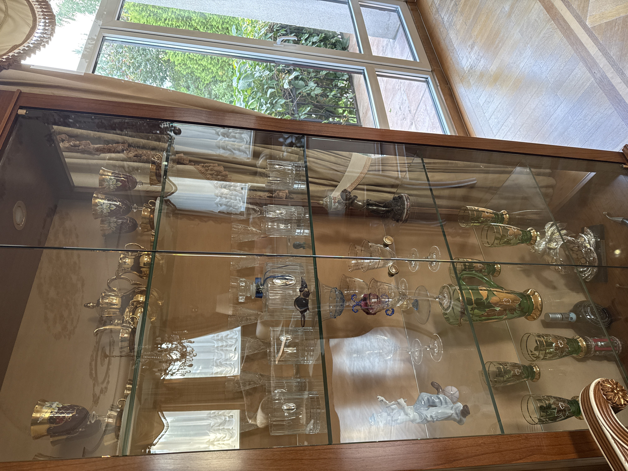 The image displays a glass-fronted display cabinet filled with various decorative items and glassware, positioned in a room with a wooden floor and large windows overlooking greenery. The cabinet is made of wood with glass shelves.
On the top shelf, there are gold-rimmed goblets and teacups, a gold teapot, a creamer, and a sugar bowl. A chandelier is visible reflected in the glass.
The second shelf from the top contains more glassware, including tall stemmed glasses and smaller cups. There is also a glass decanter with a blue ribbon.
The third shelf features several clear glass tumblers or whiskey glasses, some with metallic accents. A dark, possibly glass, object resembling a bird or animal is placed among them, and a small, decorative statue is visible on the right.
The fourth shelf holds a white ceramic figurine of a person playing a musical instrument, several clear stemmed glasses, and a green glass pitcher decorated with gold floral patterns. A dark glass bottle with a label is also present on this shelf.
The bottom shelf appears to contain more glassware and a silver decorative object.
The environment outside the window consists of lush green trees and foliage, suggesting an outdoor setting. The interior room has a warm, polished wooden floor and the overall impression is one of an elegantly furnished interior space. The location is identified as Yukki, Russia.