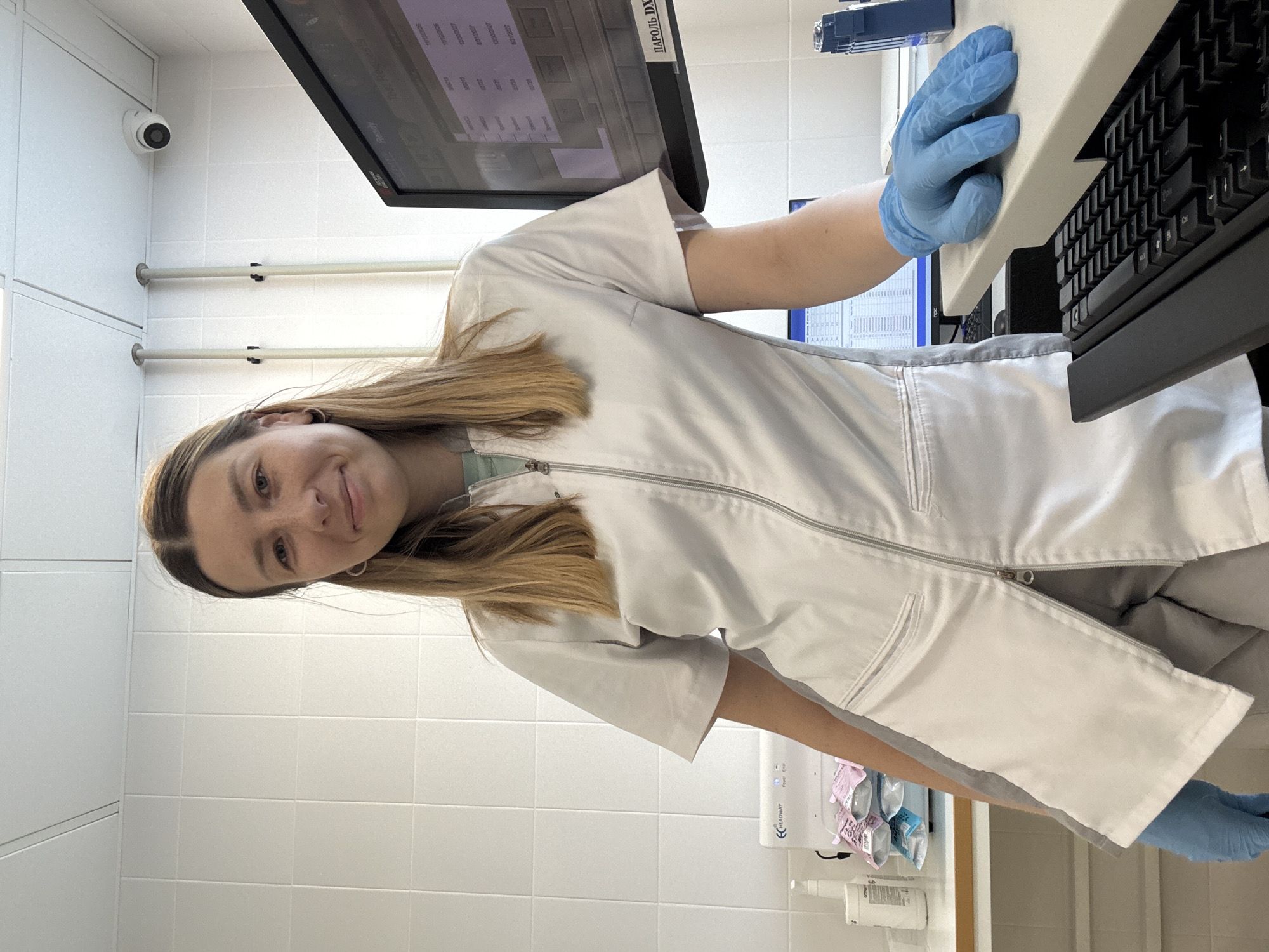 A young woman wearing a white lab coat and blue gloves stands in a laboratory. She is looking towards the camera and smiling. Her hair is long and blonde. In the background, there are white tiled walls, a computer monitor displaying data, and various laboratory equipment. A security camera is visible on the ceiling. The scene appears to be indoors, in a professional setting, possibly a medical or research laboratory. There is no indication of the specific location in Moscow, Russia.