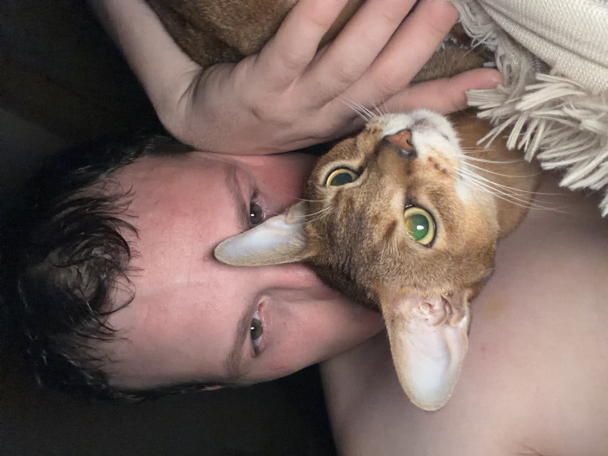 A man is lying down with a cat on top of him. The cat is reddish-brown with green eyes. The man has dark hair and light skin. The cat's ear is next to the man's face. A hand is visible behind the cat. There is a fringed blanket or cloth partially visible in the upper right. The photo does not provide enough information to determine the location as Chaikovsky, Russia.