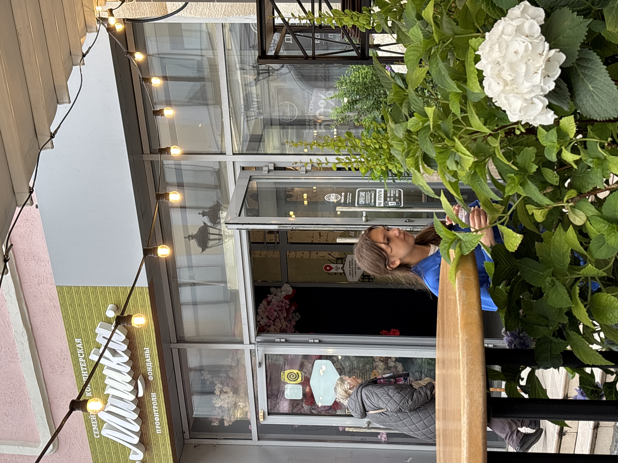 The image depicts the exterior of a cafe or confectionery shop in Nizhny Novgorod, Russia, identified by a sign reading "СЕМЕЙНАЯ КОНДИТЕРСКАЯ Манника" (Family Confectionery Mannika). String lights with illuminated bulbs are strung across the facade.
In the foreground, a round wooden table is visible, partially obscuring a young person with brown hair wearing a blue jacket, who is holding a clear cup. Beside them, a large white hydrangea bush is in bloom.
To the left, an older woman with gray hair, wearing a dark quilted jacket, is walking towards the entrance of the establishment. The entrance features glass doors, one of which is ajar, revealing floral decorations inside. A sign on the door reads "COFFEE CAKE" and displays operating hours.
Reflected in the glass doors are images of lamps and possibly other shops. Further inside the establishment, a sign with "50%" discount information is visible on the window. The overall environment suggests a casual outdoor seating area of a commercial establishment.