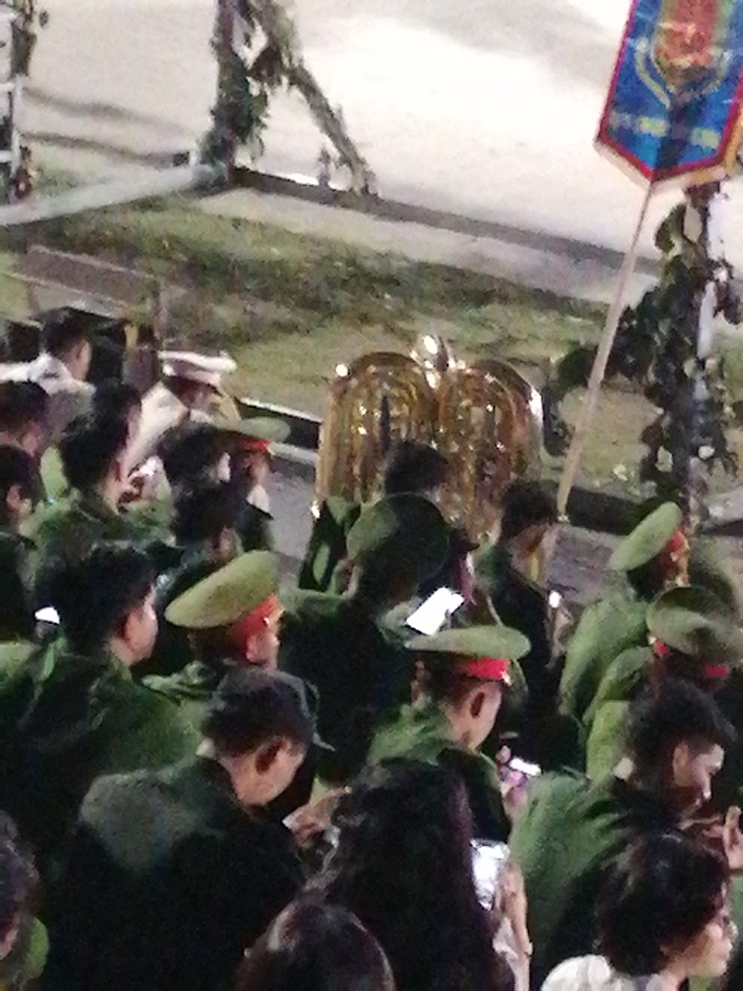 The image depicts a crowd of people, many wearing green uniforms and hats, suggesting a formal event or procession. The people are gathered outdoors, with what appears to be a paved surface and some grassy areas visible. In the background, there's a glimpse of foliage and what might be a structure. A flag with blue and red colors, possibly a ceremonial banner, is held on a pole. The lighting suggests it might be evening or overcast. The overall scene conveys a sense of public gathering.