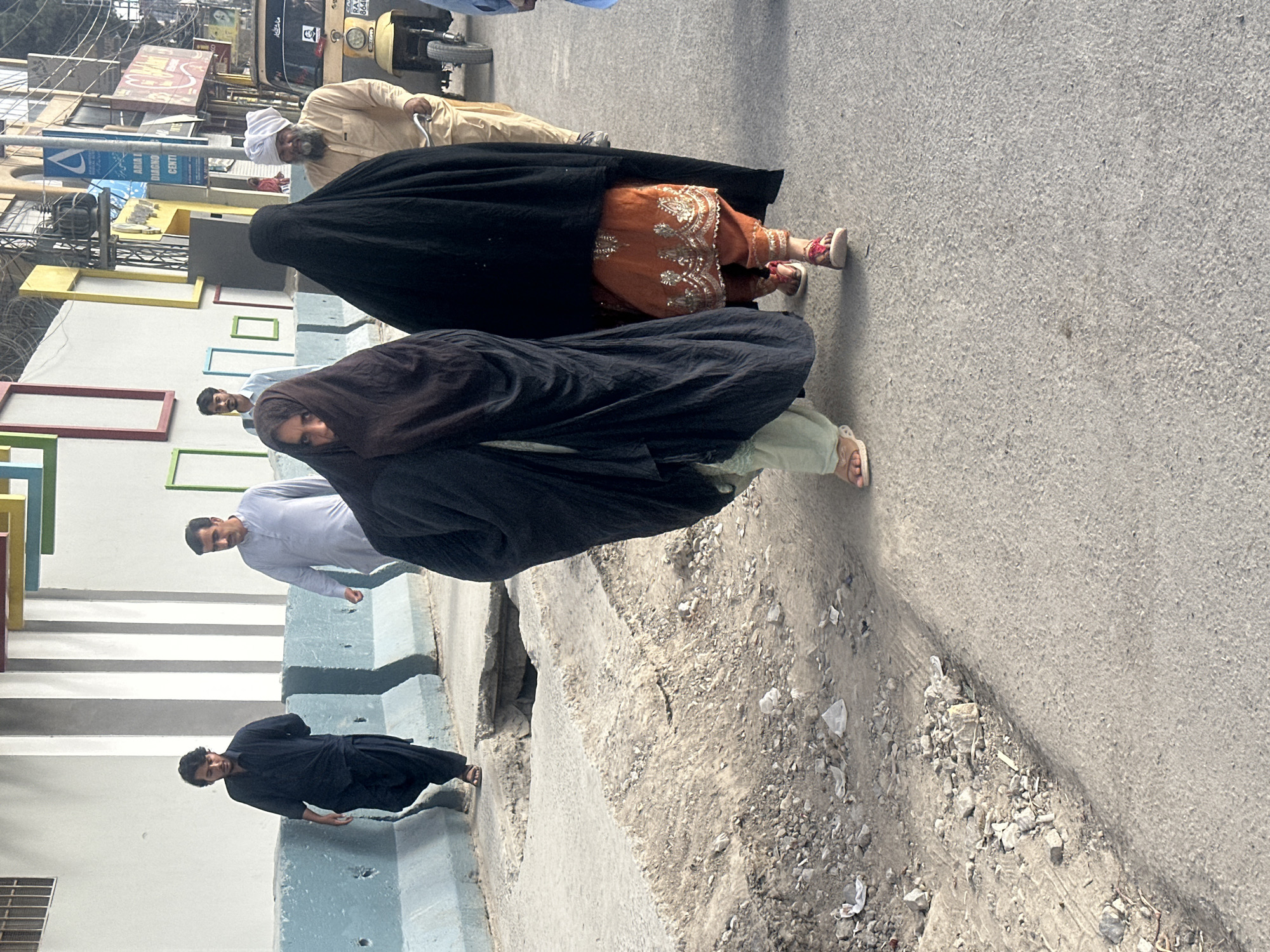 In Quetta City Tehsil, Pakistan, a group of individuals walks along a city street. Two women, each covered in a dark burqa, are in the foreground, walking closely together. The woman on the right is wearing an orange shalwar kameez with intricate silver embroidery. Behind them, a man with a white turban and beard, dressed in a tan shalwar kameez, walks beside a three-wheeled auto rickshaw. Further back, two men in traditional shalwar kameez are walking in the same direction. One man is wearing a light blue shirt and the other a dark blue shirt. The street is paved and appears to be under some disrepair with debris on the side. The background shows buildings with colorful decorative elements and street signs.