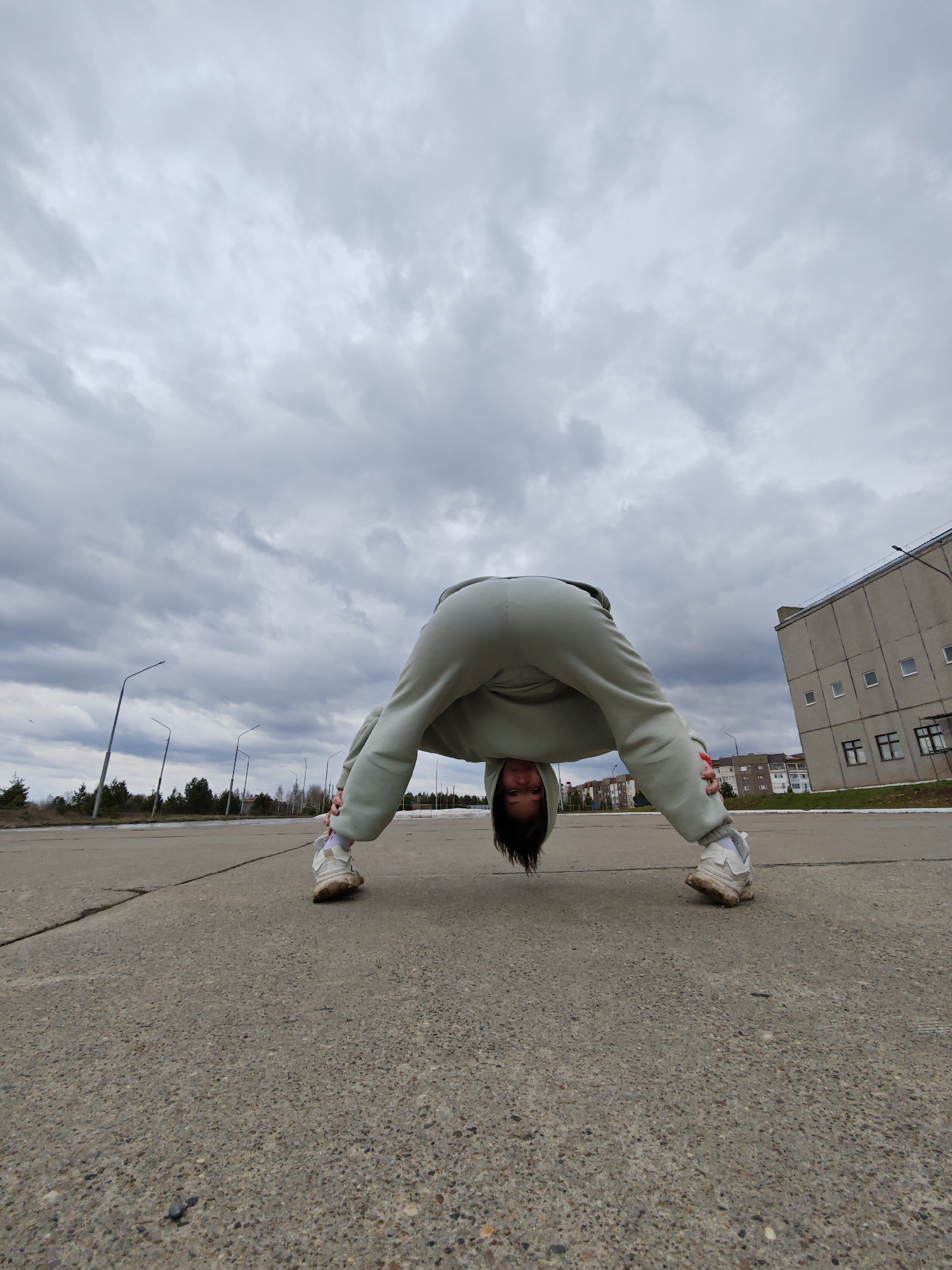 The image depicts a person performing a wide-legged forward fold or stretch on a paved outdoor surface in Markovskii, Russia. The individual, seen from a very low angle, is dressed in a light green or mint-colored sweatsuit, comprising a hooded top and sweatpants, worn with white, somewhat dirty sneakers. Their head hangs upside down between their spread legs, with dark hair falling towards the ground, and they look directly at the camera with a neutral to slightly smiling expression.
The setting is an expansive, cracked asphalt or concrete area. In the background, under an overcast sky filled with grey clouds, streetlights line what appears to be a road. Beyond the road, a strip of trees, including both coniferous and bare deciduous trees, separates the foreground from the distant urban landscape. To the right, a large, grey, multi-story building with small rectangular windows suggests industrial or older institutional architecture. Further back, residential buildings are visible in the distance.
The weather is cloudy and grey, indicating daytime without direct sunlight, possibly in early spring or late autumn given the presence of bare trees. No visible text is present in the image. The scene conveys an active and somewhat playful moment against a stark, urban-natural backdrop.