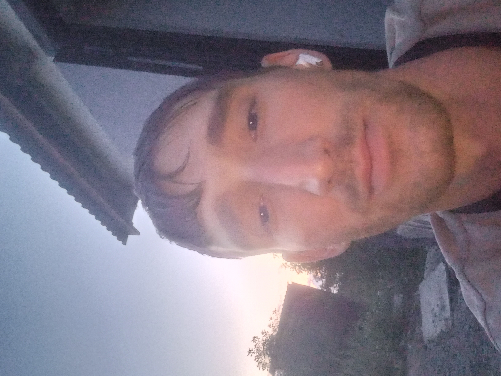 A medium shot selfie is taken from a slightly low angle by a fair-skinned man with short, dark hair and stubble. He is looking directly at the camera with a neutral expression. The man is wearing a light-colored t-shirt and has a white wireless earbud in his right ear. In the background, there is a partial view of a building with a corrugated roof. The setting appears to be outdoors, with trees and vegetation visible to the left, suggesting a rural or semi-rural environment. The sky in the background is a pale, dusky blue with a hint of pink, indicating either early morning or late evening. The location is identified as Қозыкеткен ауылдық округі, Kazakhstan. The overall lighting is dim, consistent with twilight hours.