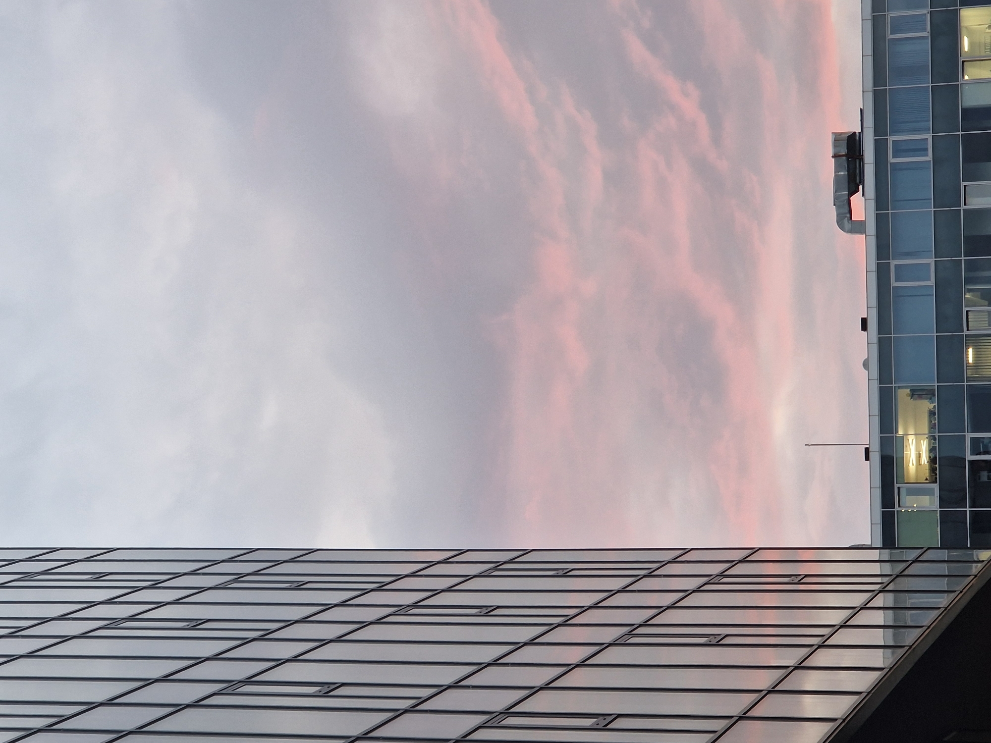 The image, when viewed in its correct orientation (rotated 90 degrees counter-clockwise), captures an urban sky at either sunrise or sunset, framed by two modern buildings. The upper and central areas are filled with a soft expanse of clouds, displaying delicate shades of pink and rose blended with light grey and white.
On the right side of the frame, a multi-story building features a facade of square and rectangular windows, some of which reflect the colored sky, while others show interior lights turned on, suggesting dawn or dusk. A large, grey ventilation duct or pipe system is visible on the upper part of this building, along with a slender antenna or pole.
The lower-left section of the image is occupied by the reflective, angled roof or facade of another modern building. This structure is composed of numerous rectangular glass panels arranged in a precise grid, reflecting the sky above it. The clean, geometric lines of the architecture provide a stark contrast to the organic forms and colors of the sky. There are no people or discernible activities taking place, and no visible text. The scene conveys a tranquil, abstract urban aesthetic.