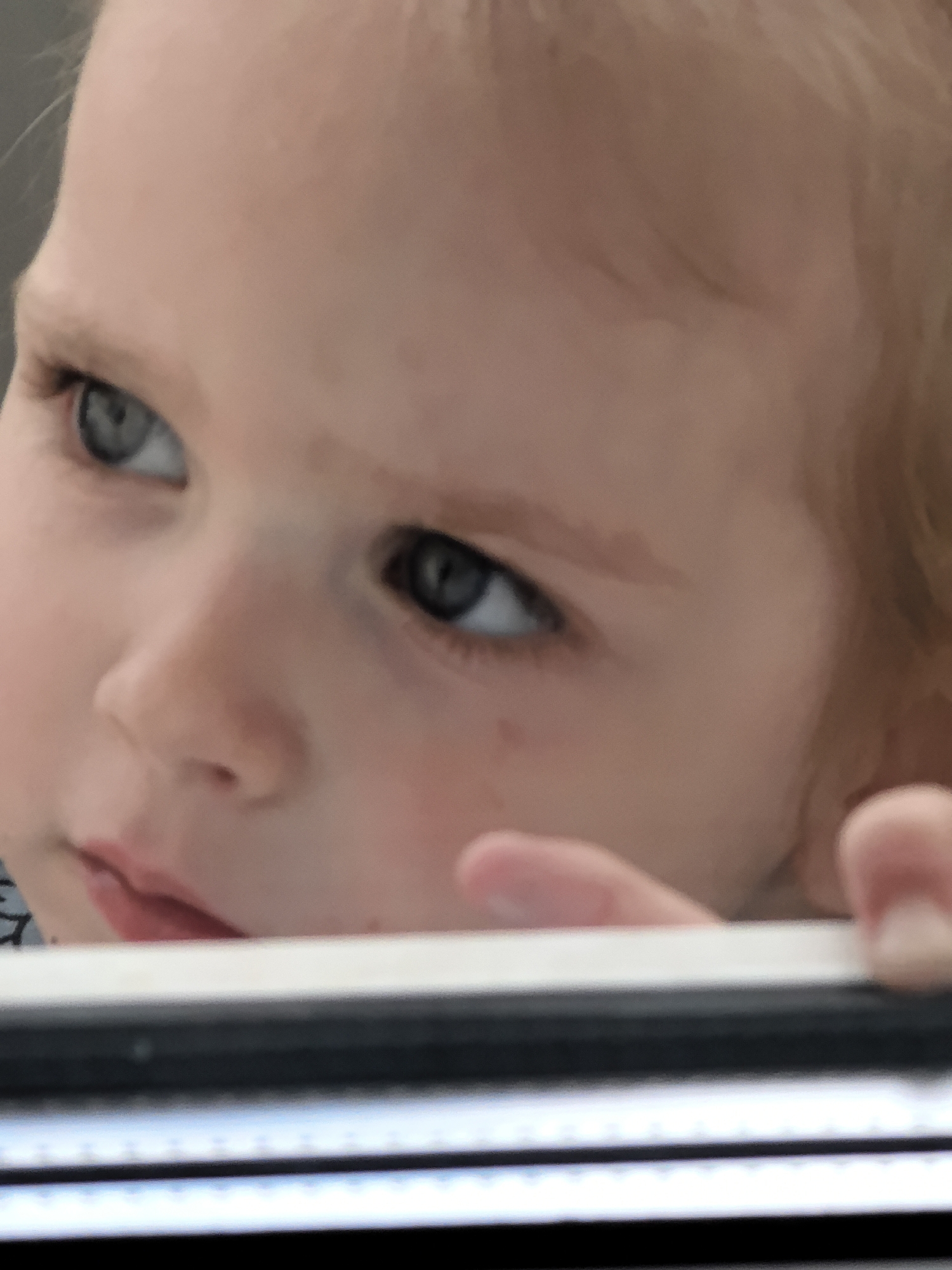 A close-up, outdoor, eye-level shot shows the face of a young child. The child is looking to their left. The child has light-colored hair, and their eyes are blueish-gray. The child's skin is fair. The child's mouth is slightly open. Part of the child's forehead and nose are visible. The child's left hand is visible near their face. A white, rectangular object is in the foreground of the image, obscuring the bottom of the child's face. The background is out of focus and appears to be a gray, outdoor environment.