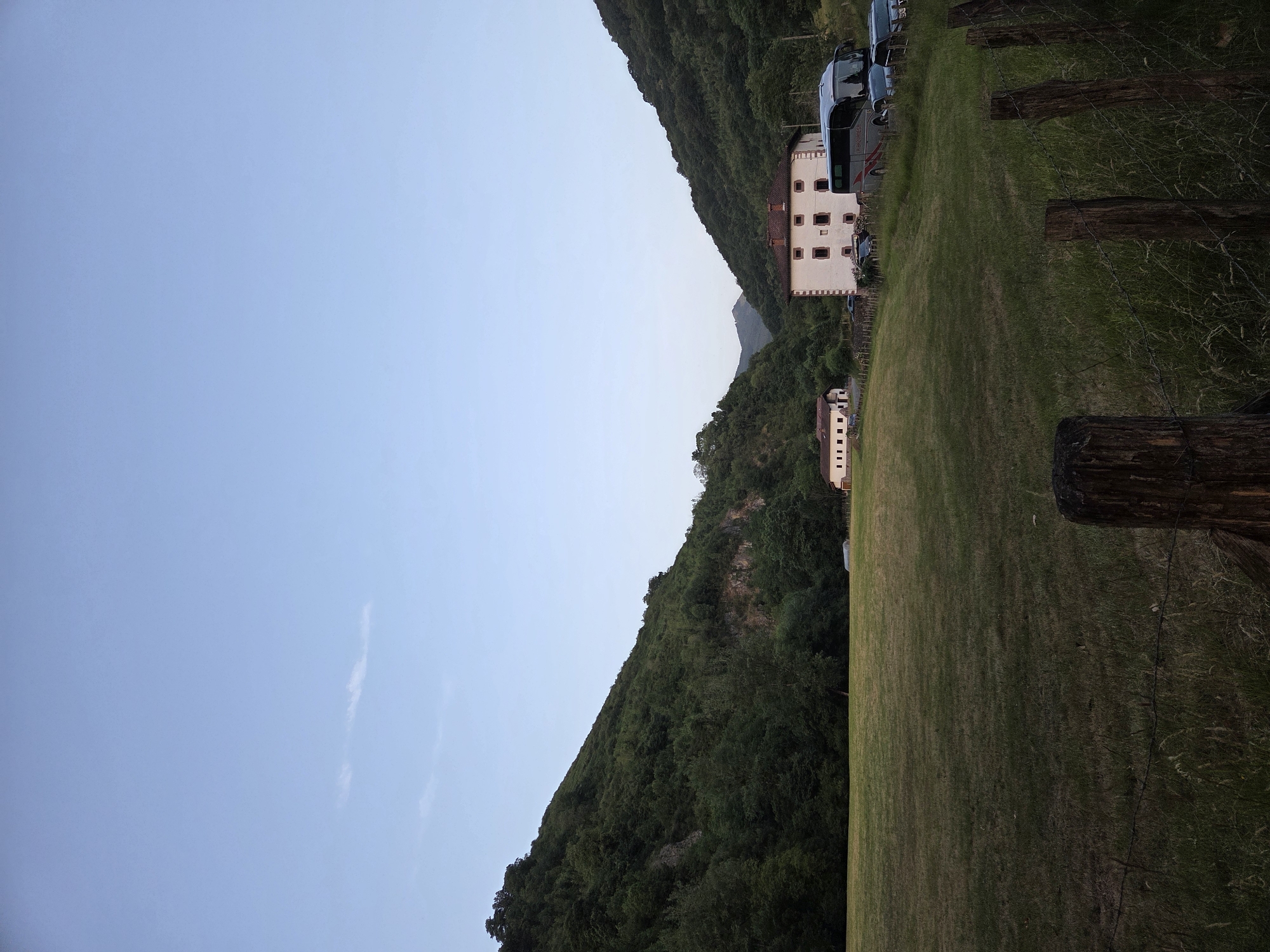 The image depicts a rural landscape in the city of Baztan, Spain. In the foreground, a wire fence with wooden posts marks the edge of a grassy field. The field itself is mostly dry and textured, with taller grass and weeds growing closer to the fence. Beyond the field, a white building with a red-tiled roof stands prominently. This building has multiple windows with dark shutters and appears to be a substantial structure, possibly a guesthouse or hotel, indicated by a tour bus and several parked cars in front of it. To the left of this building, a smaller, white building with a similar red-tiled roof is visible, suggesting a complex of structures. The backdrop consists of two large, densely forested hills that create a valley. The sky is clear with a faint gradient of blue, hinting at either early morning or late evening. The overall scene conveys a sense of tranquility and natural beauty.