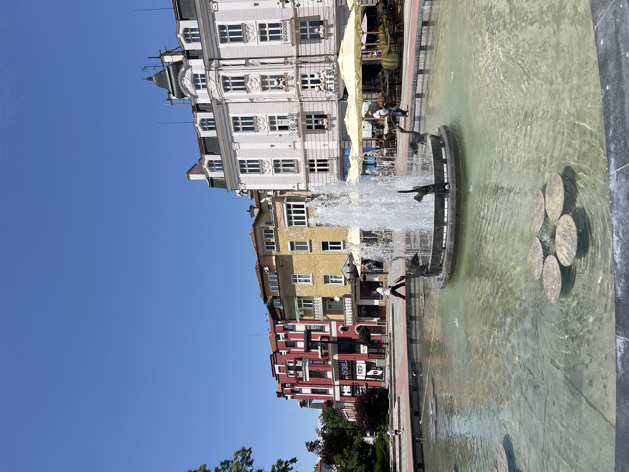 The image captures a street scene in Plovdiv, Bulgaria, showcasing a water fountain situated in an urban square. Buildings with varied architectural styles, painted in red, yellow, and white, line the perimeter of the square. A fountain with multiple levels and decorative dolphin statues is centrally positioned, its water spray creating a visual focal point. A person is standing near the fountain and another walks in the background. Cafe seating and parasols are seen near the white building. The sky is clear and blue.