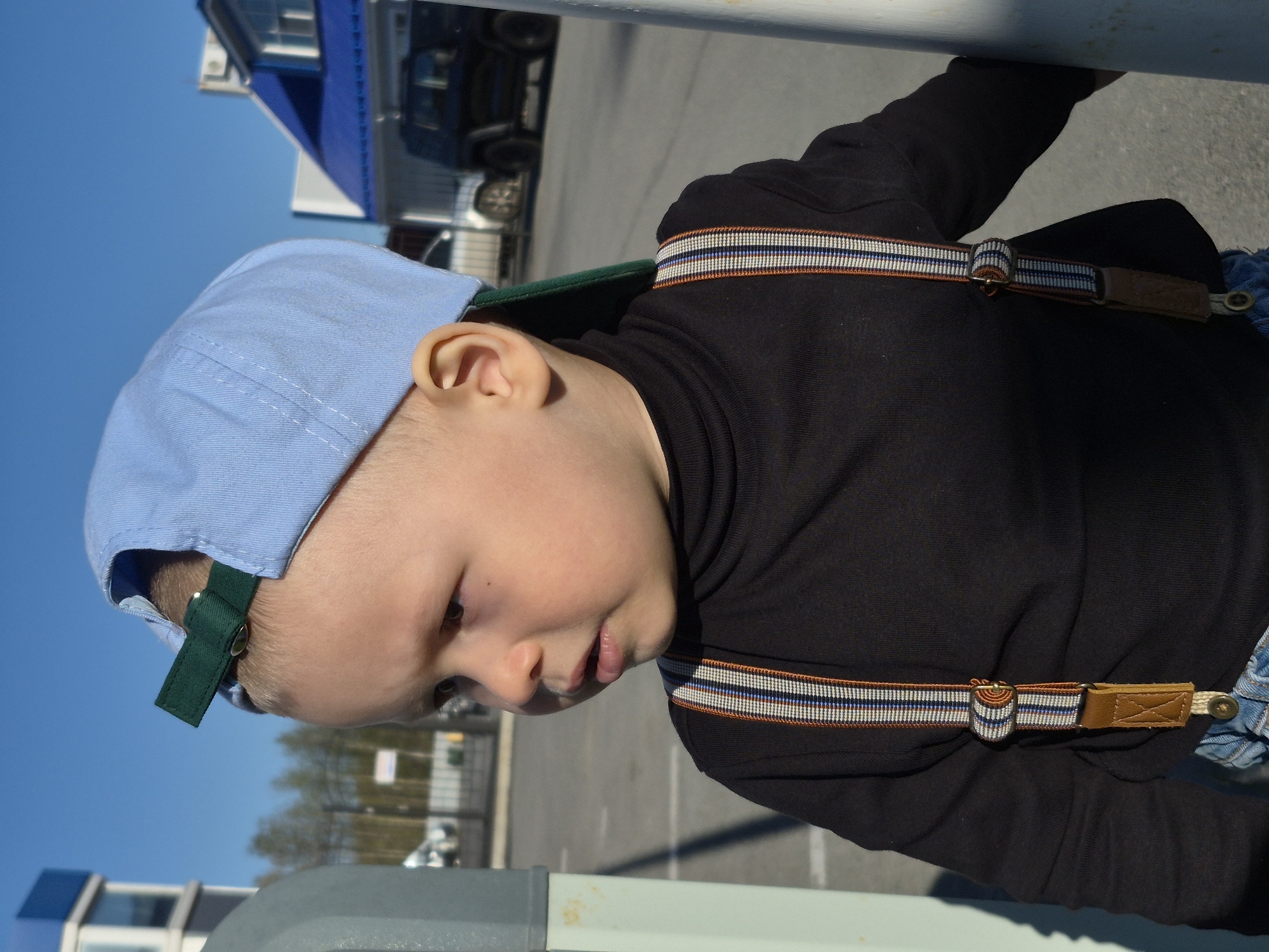 A young child wearing a blue cap, black turtleneck, and suspenders is standing outside on a sunny day. The background includes a building, vehicles, trees, and a clear blue sky. The location is suggested to be in Tobolsk, Russia, based on provided information.