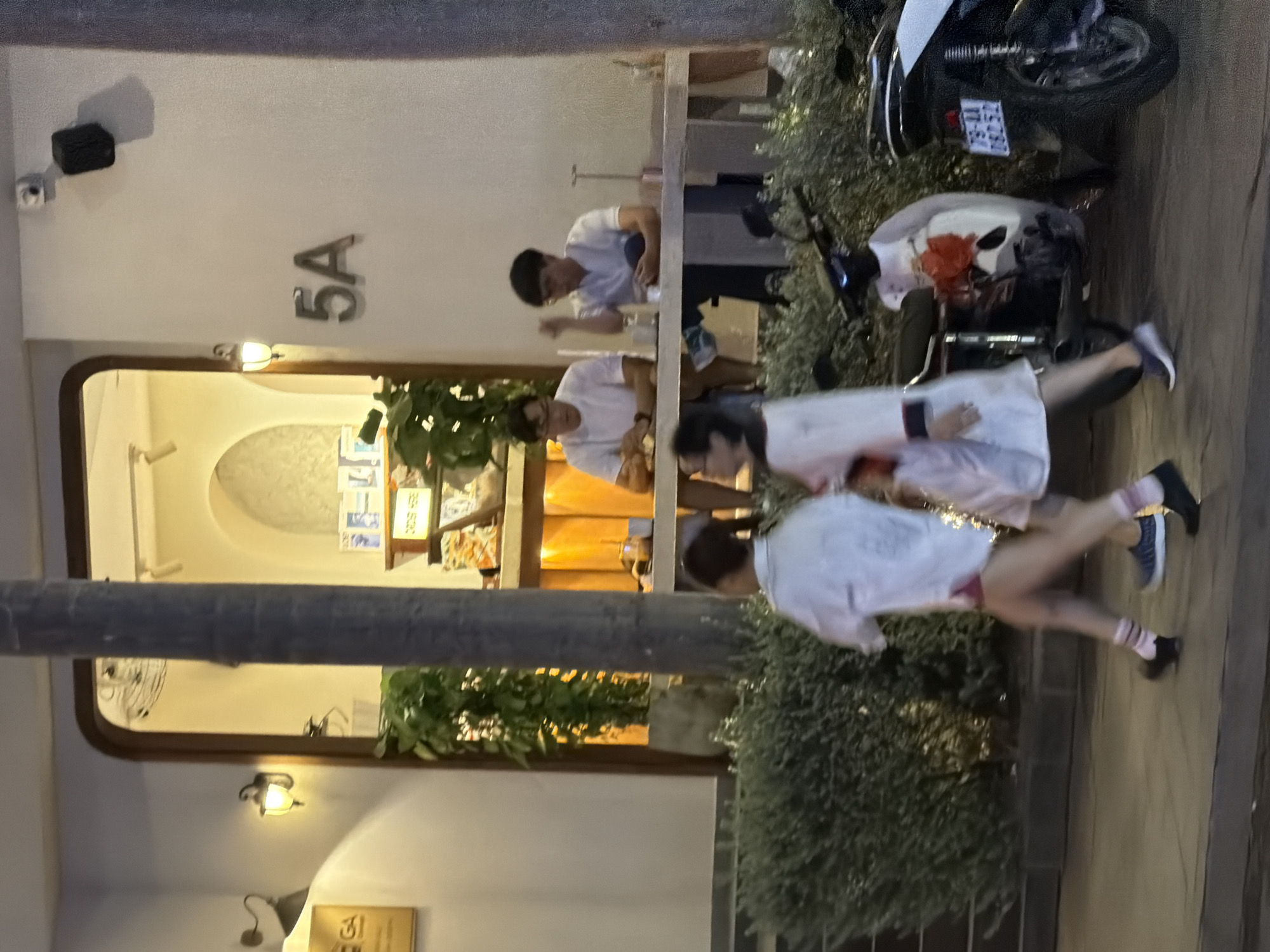 The image depicts a street scene at dusk in Nha Trang, Vietnam. Two individuals, a man and a woman, are walking from left to right in the foreground, their legs and lower torsos visible as they move across a paved sidewalk. They are wearing casual clothing, including socks and athletic shoes. Behind them, a scooter with floral decorations is parked.
In the background, a building with a wide, arched entryway is illuminated by warm interior lighting. Two men are seated at a table within the building, appearing to be engaged in conversation or a meal. One man wears glasses and a white t-shirt, while the other also wears a white t-shirt. The building's facade features the number "5A" prominently displayed. Further to the left, a sign with the letters "GA" is visible on the building's exterior. Lush green bushes line the front of the building. The overall lighting suggests it is evening.