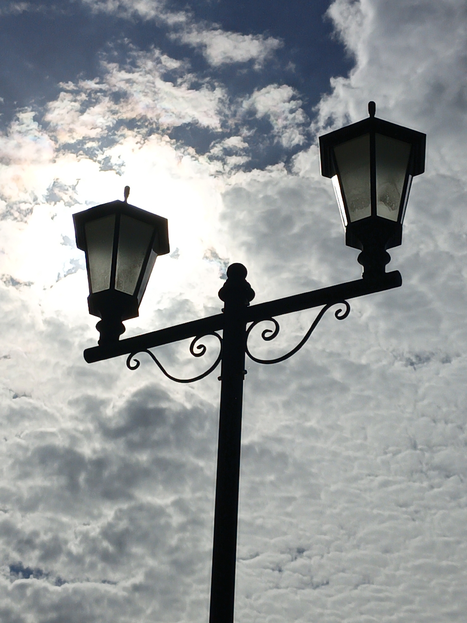The image shows a black silhouette of a double lamppost against a cloudy sky. The lamppost has a central pole, a horizontal arm with decorative swirls, and two lantern fixtures with peaked roofs. The lanterns appear to be illuminated. The sky is filled with white and gray clouds, with the sun partially obscured behind the clouds, creating a bright area behind the left-hand lantern. The image was taken in the city of Балаклавский округ, Ukraine.