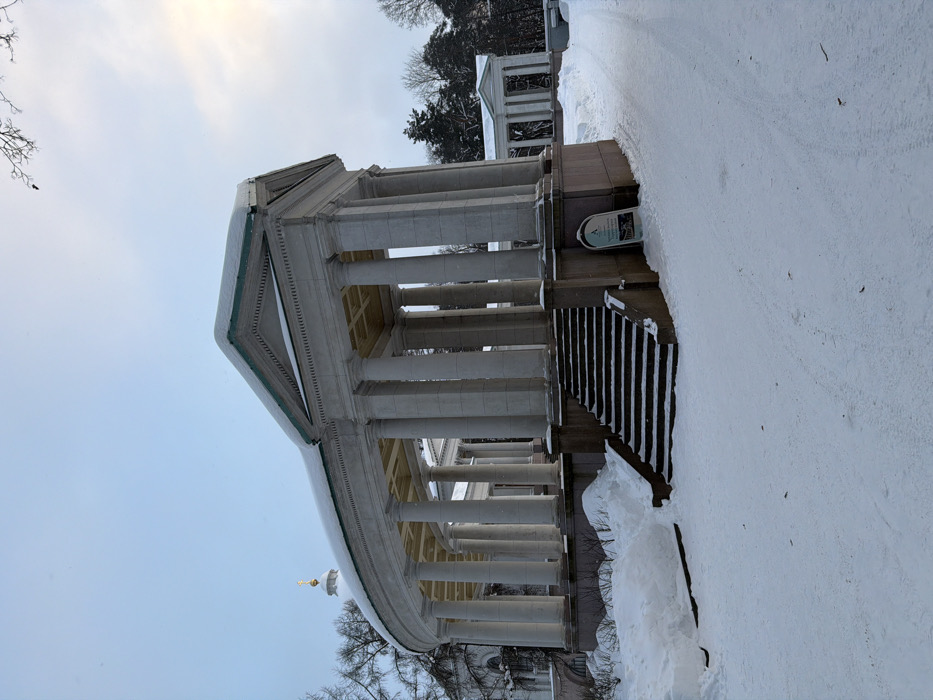 The image displays a snow-covered neoclassical colonnade in посёлок дачного хозяйства «Архангельское», Russia, under an overcast light blue sky, brighter towards the upper right. The central subject is a large, light-colored curved colonnade, featuring numerous columns supporting a roof, with a straight end section topped by a pediment on the right. A thick layer of white snow covers the roof, revealing a green trim along its edge. Dark-colored steps, partially obscured by snow, ascend to the raised base of the colonnade. The ground is uniformly covered in snow, with significant drifts accumulated to the left of the stairs. A snow-cleared path extends to the right, adjacent to which stands a light-colored A-frame sign with dark lettering and a logo. In the background, a white-domed building with a golden, cross-topped spire is visible behind the left curve of the colonnade, and another smaller columnar structure is present further right amidst snow-dusted bare and coniferous trees. No individuals are present.