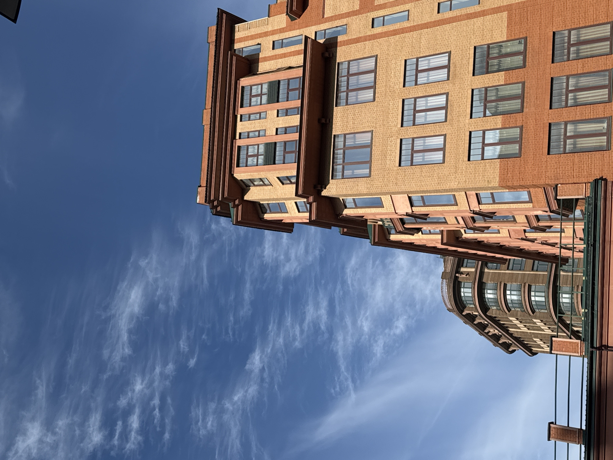 The image shows two buildings against a blue sky in Saint Petersburg, Russia. The buildings are brick with many windows. The sky has some wispy clouds. The photo is taken from a low angle.