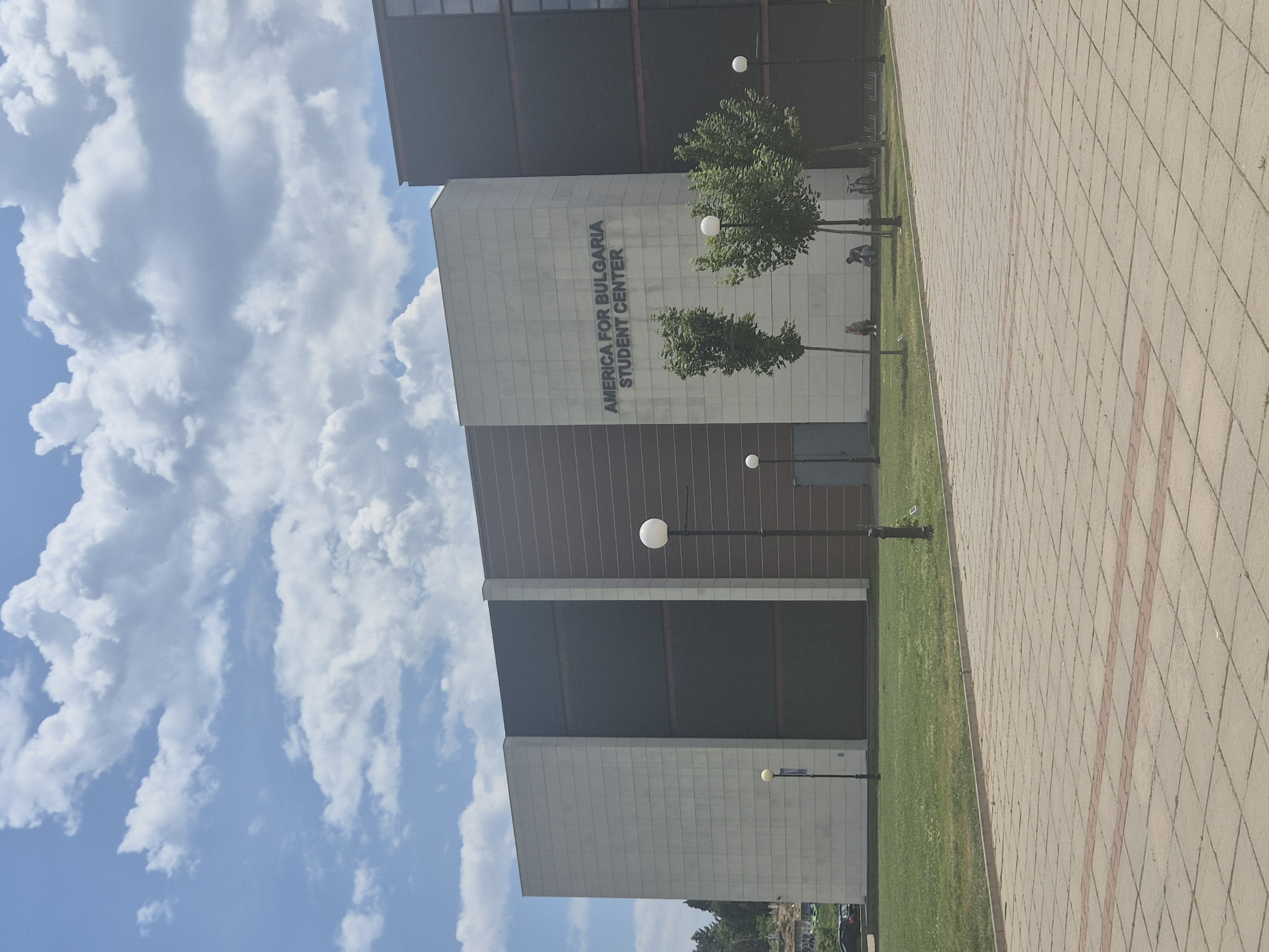 The image depicts the "America for Bulgaria Student Center" building, a modern structure with a mix of grey, brown, and black panels, located in Blagoevgrad, Bulgaria. The building is surrounded by a neatly maintained lawn and a paved walkway made of tan and reddish bricks. There are small trees and globe-shaped streetlights spaced along the walkway. The sky is bright blue with significant cloud cover. Two people are faintly visible on the sidewalk by the trees.
