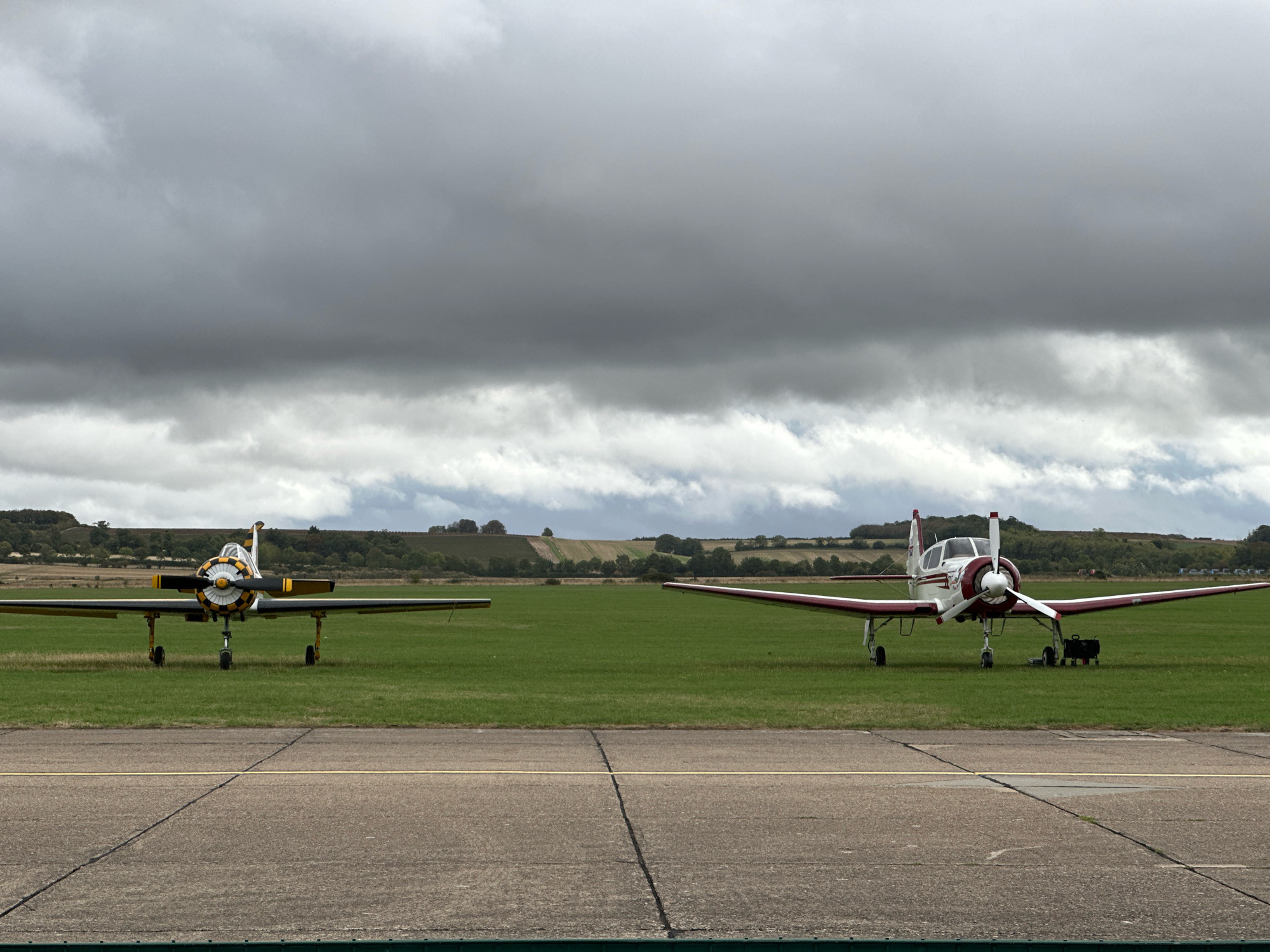 Two light aircraft are parked on a grassy airfield. The aircraft on the left is yellow and black with a propeller. The aircraft on the right is white and red with a propeller. Both aircraft are facing forward. The airfield is located in Cambridge, United Kingdom, under a cloudy sky. The foreground shows a tarmac runway with yellow and white markings. In the background, there are rolling hills with trees and fields.