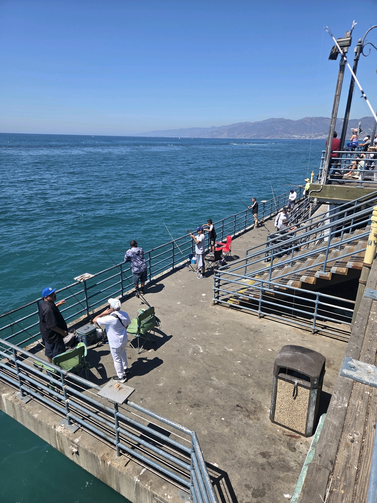 This image captures a sunny day on the Santa Monica Pier in Santa Monica, California. The scene shows a section of the pier with a clear blue sky overhead and the vast, turquoise ocean stretching out to the horizon. Several people are present, engaged in fishing. Two men in the foreground are standing near the railing, one wearing a blue cap and black shirt, the other a white shirt and pants. Between them are green folding chairs and a cooler. Further along the pier, other individuals are also seen fishing, holding rods and casting lines into the water. The pier itself is constructed with wooden planks and has metal railings. A set of stairs leads up to a higher level where more people are gathered. A dark-colored trash receptacle with a granular filling is visible on the pier surface. In the background, mountains are visible on the coastline.