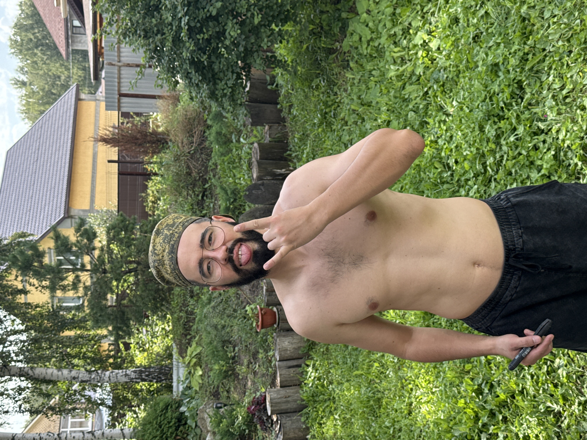 A shirtless man with a beard and glasses stands in a grassy backyard. He wears a camouflage patterned skullcap and dark sweatpants. He is sticking his tongue out and holding his hand up in a rock-and-roll gesture. He is holding a black pen in his other hand. In the background, there are two houses and a wooden fence. The location is identified as Kremenkul', Russia.