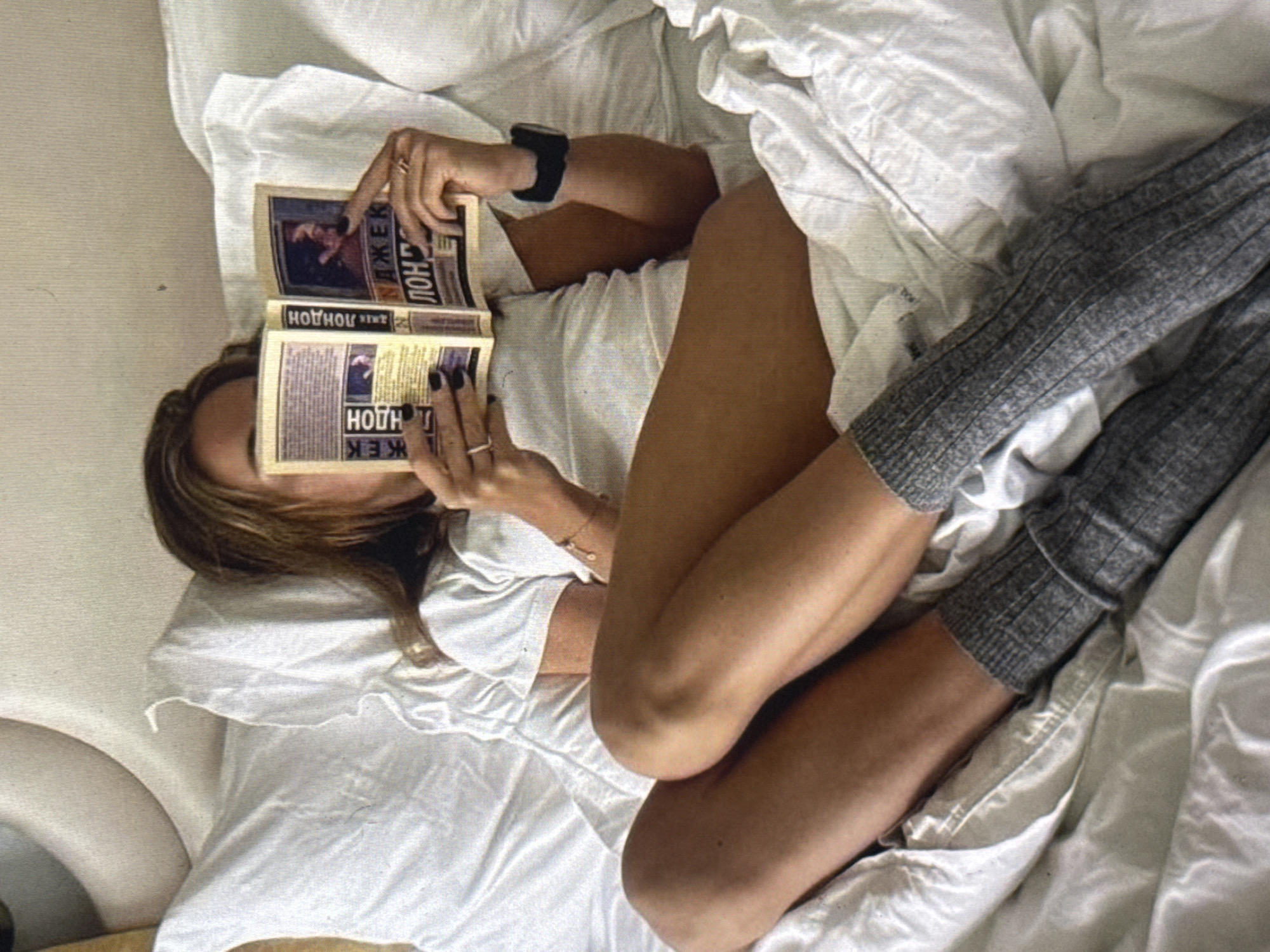 The image depicts a person lying in bed, holding two open newspapers. The individual is partially covered by white bedding and wears gray, ribbed socks. Their legs are crossed, and one arm is raised, holding a newspaper to their face, obscuring their features. The other hand holds a second newspaper. The scene appears to be indoors, in a bedroom. The lighting is soft, suggesting it might be daytime or a dimly lit room. The overall impression is one of relaxation and reading. Visible text on the newspapers includes what appears to be Cyrillic script, with some letters resembling "Н," "О," "Л," and "Ж." Due to the context provided, the location is Kuta Selatan, Indonesia.